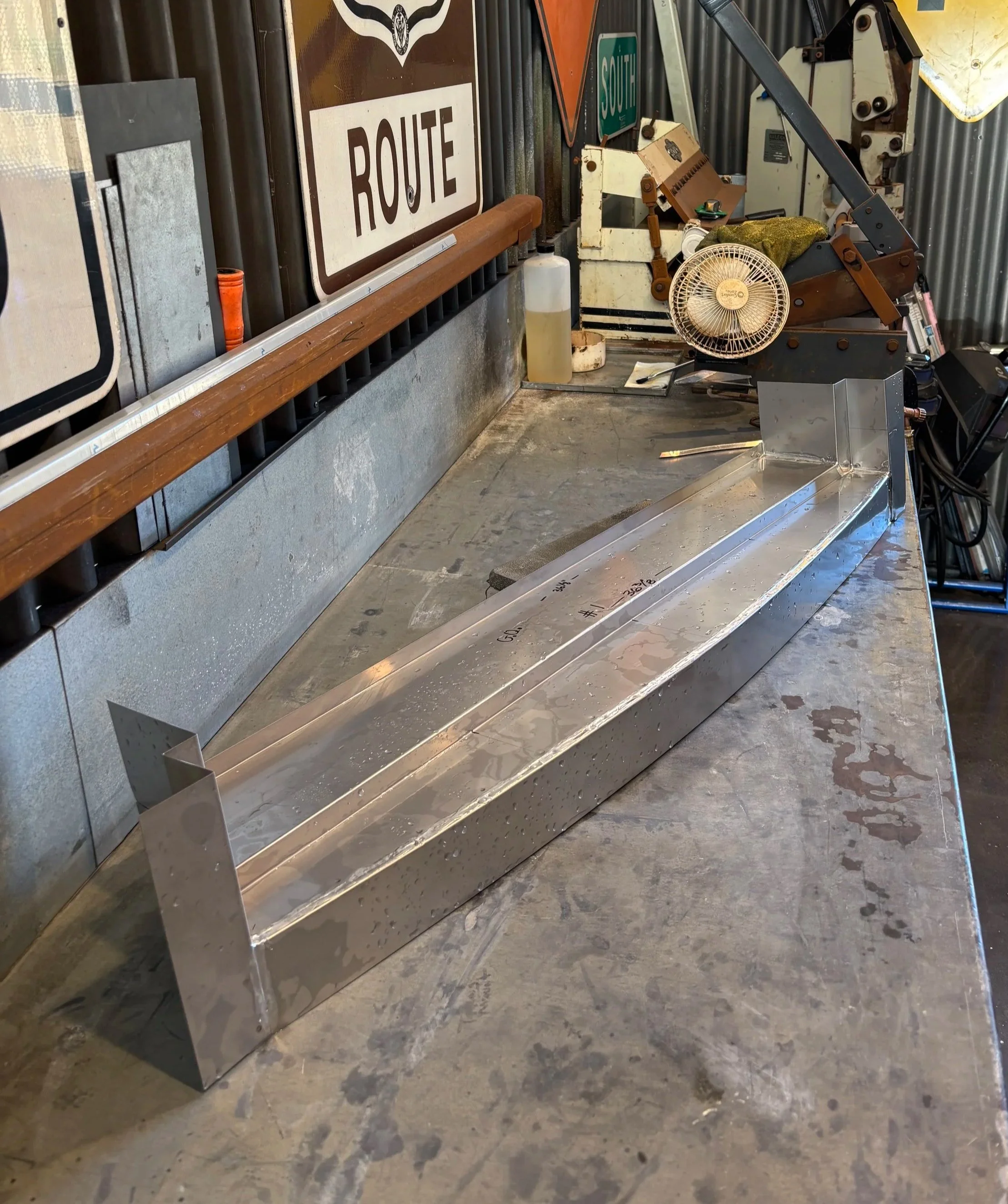 Metal workbench with various tools and objects including a small fan, a plastic bottle, and a long metal piece. Behind, there are signs on a metal wall, one with the word 'ROUTE' and another with 'SOUTH.'