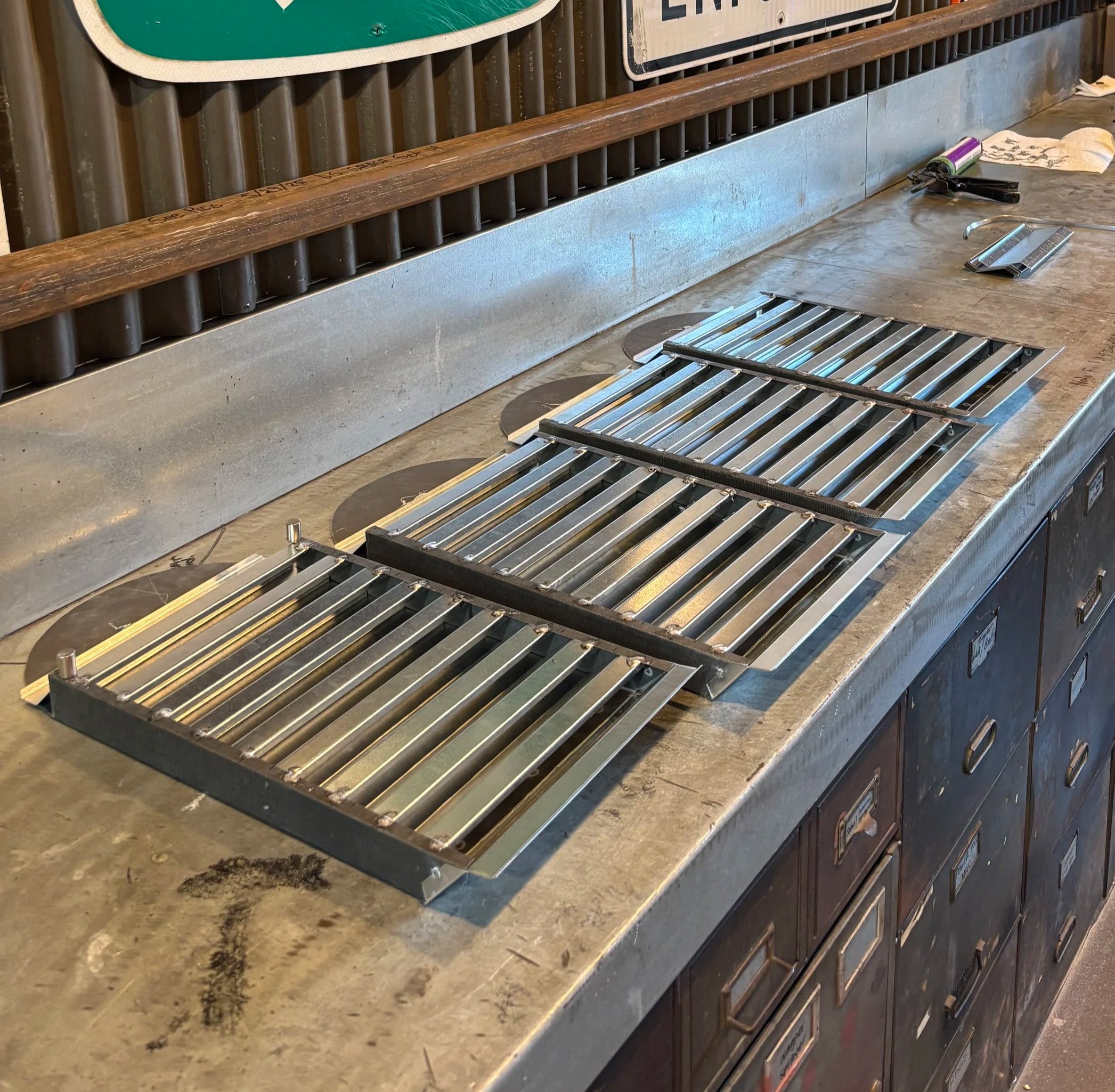 Metal and wood construction tools on a workbench, including a grater, a can of spray paint, and metal bars, with a vent grate on the floor and part of a sign in the background.