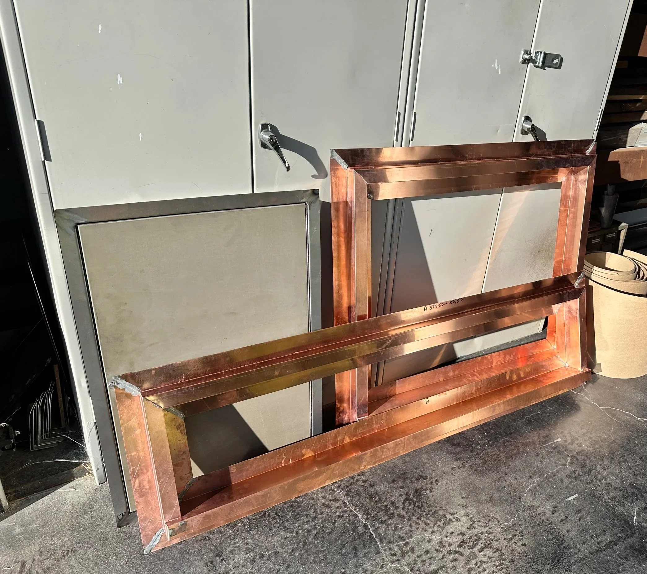 Metal lockers in beige, with two handles and a latch, set against a black marble floor, with parts of a wooden frame and a black wire shelf nearby.
