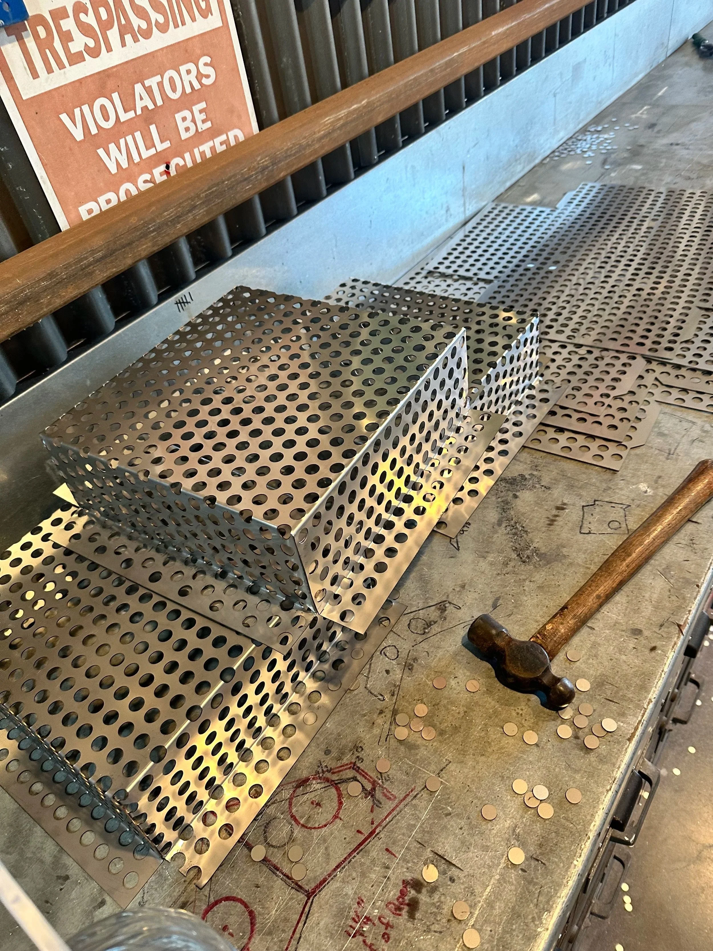 Metal perforated sheets laid out on a workbench with scattered coins, a hammer, and a warning sign in the background that reads 'Trespassing Violators Will Be Prosecuted'.