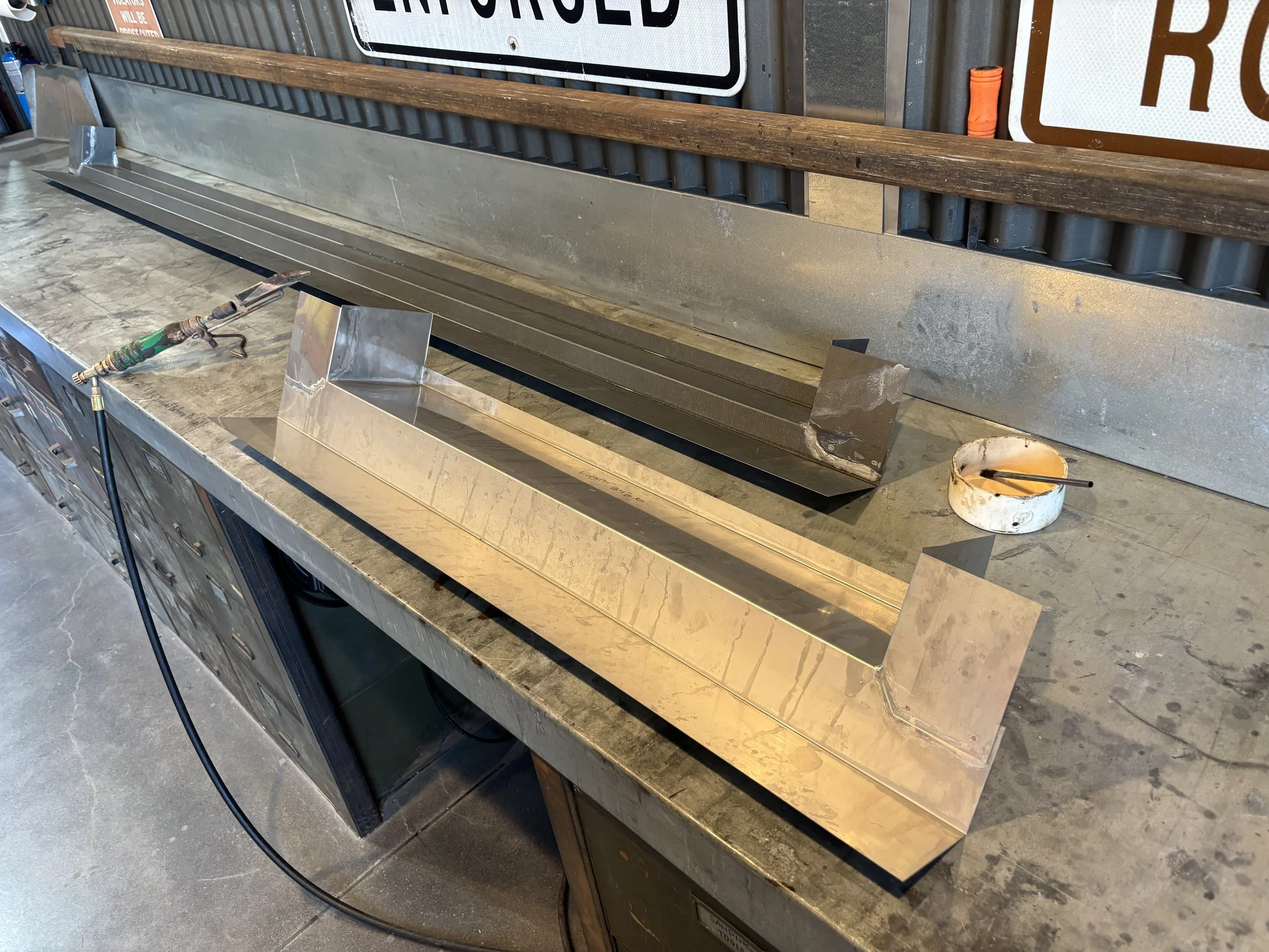 Workshop workspace with a metal workbench, welding tools, a bucket with liquid, signs, and a wooden rail mounted on a wall.