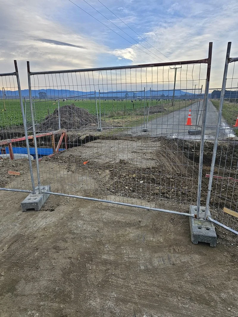 Construction site on a dirt path surrounded by a metal fence, with traffic cones, in a rural area with green fields and mountains in the background under cloudy sky.