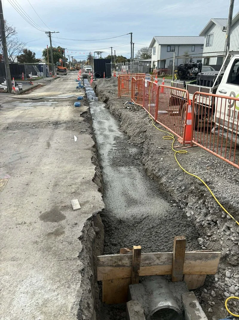 Construction site on a street with a long trench being dug for underground utility work, with orange safety barriers and construction equipment.