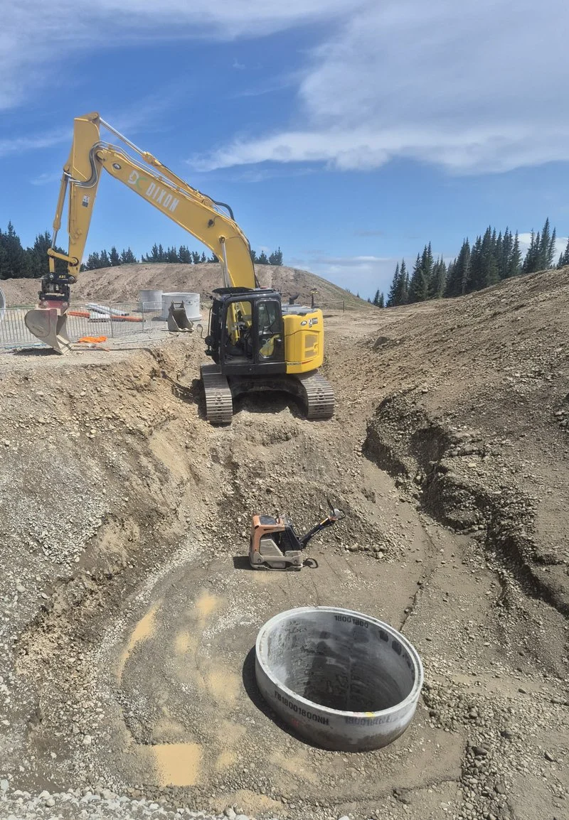 Construction site with a yellow excavator working on a large hole, nearby a concrete pipe and construction tools, under a blue sky with scattered clouds.