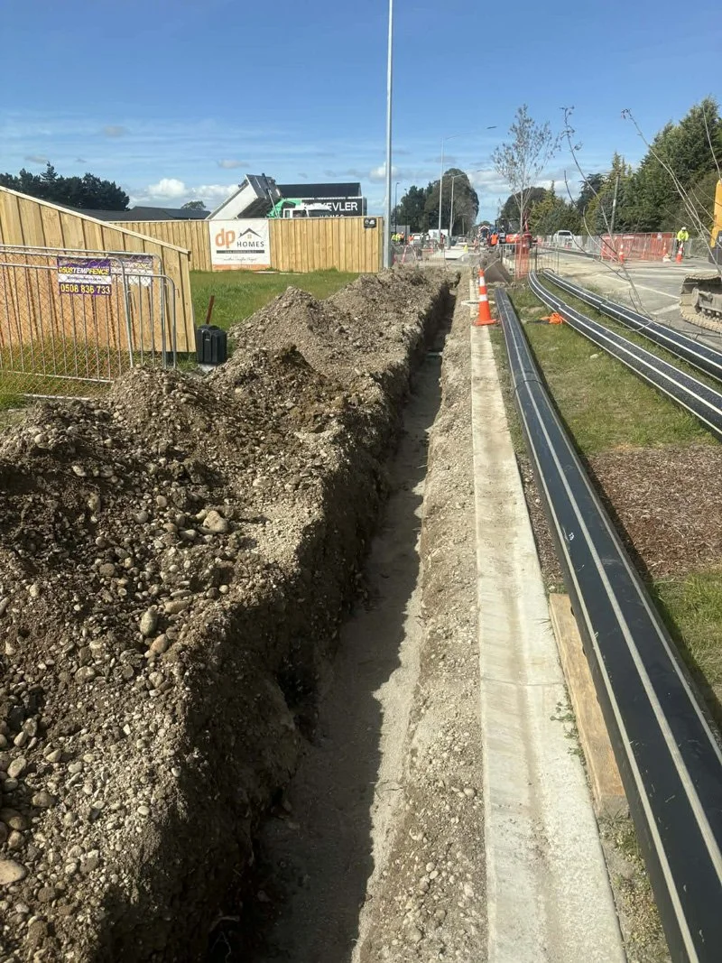 Construction site with a trench dug alongside a sidewalk, orange traffic cones, and a partially installed metal railing.