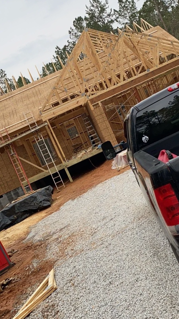 Construction site of a house with wooden framing, scaffolding, and a truck parked nearby in a wooded area.