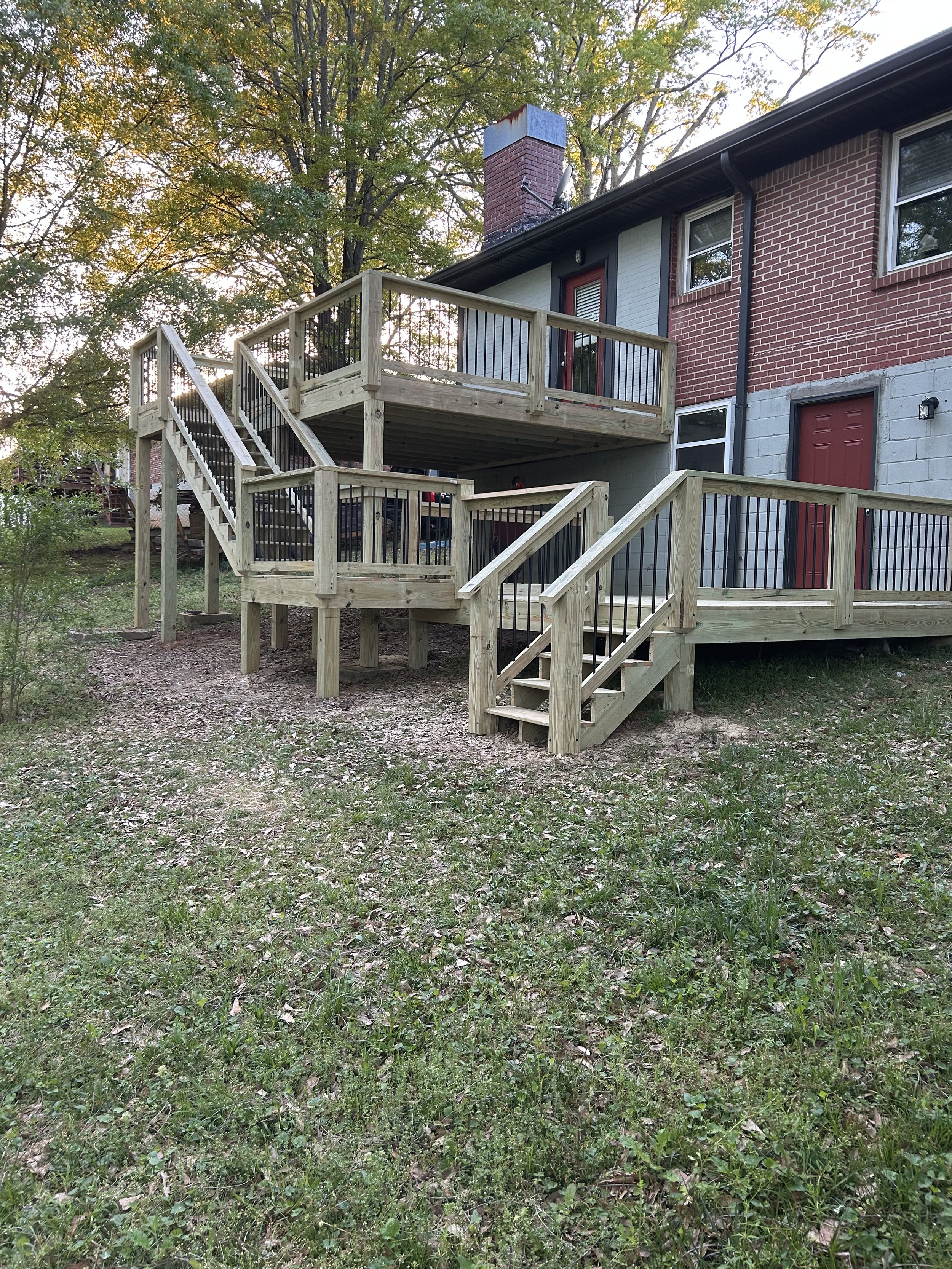 Newly built wooden deck with stairs attached to the back of a brick house with large windows and a chimney, surrounded by green trees and grass.