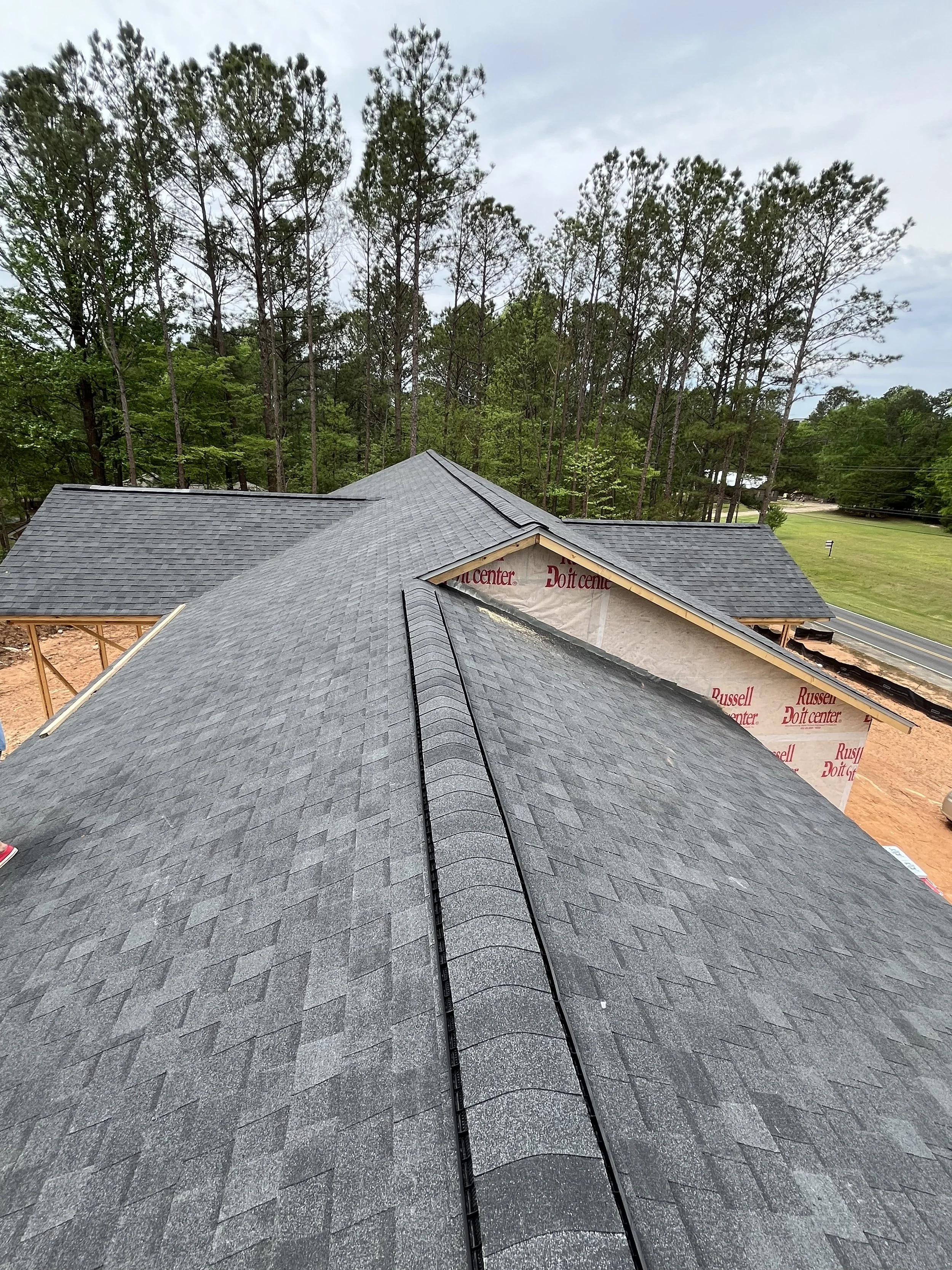 View of a house roof under construction with asphalt shingles, surrounded by trees and a partly cloudy sky.