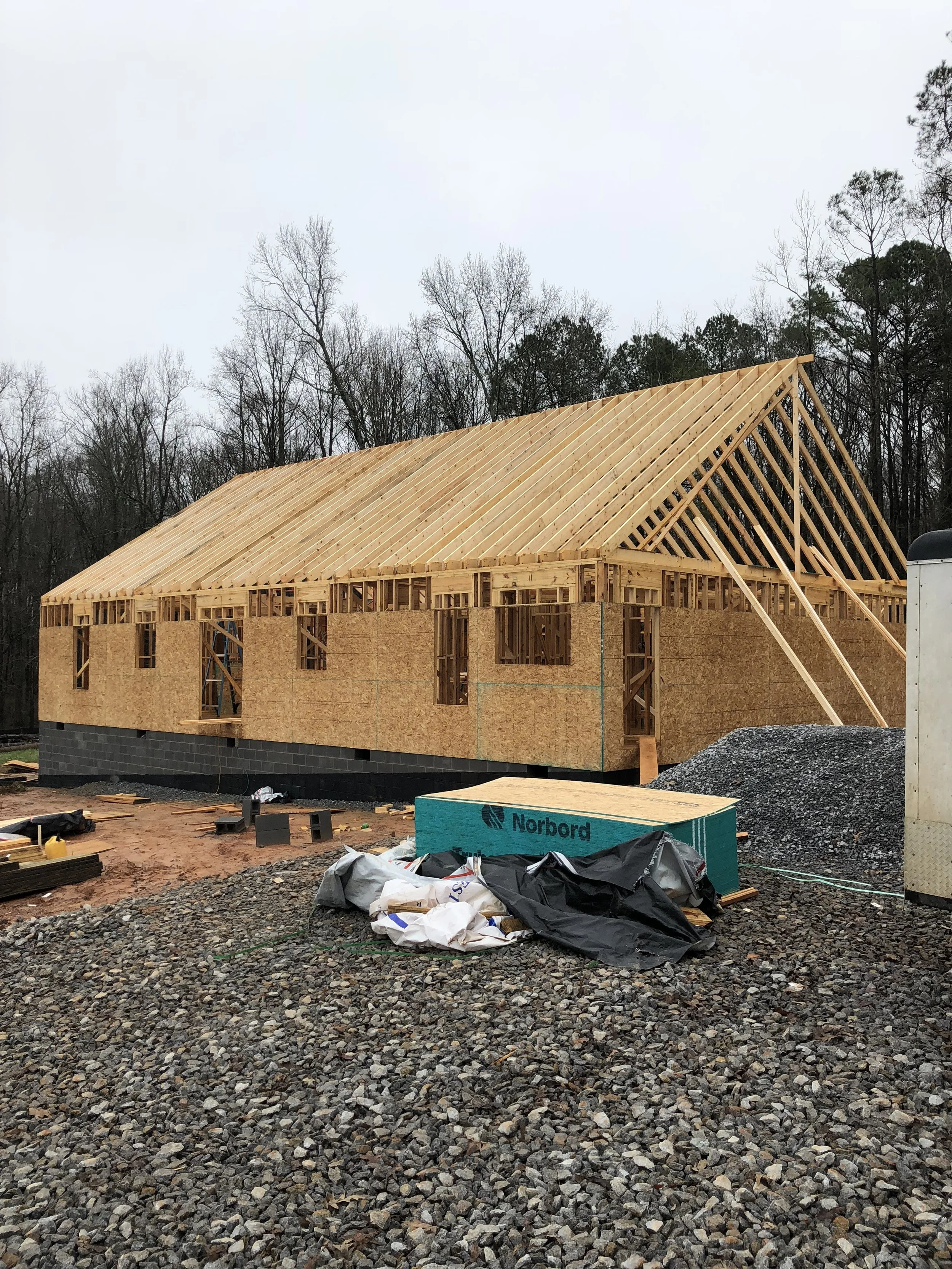Construction site of a house with wooden framing and a roof structure in progress, surrounded by gravel and construction materials.