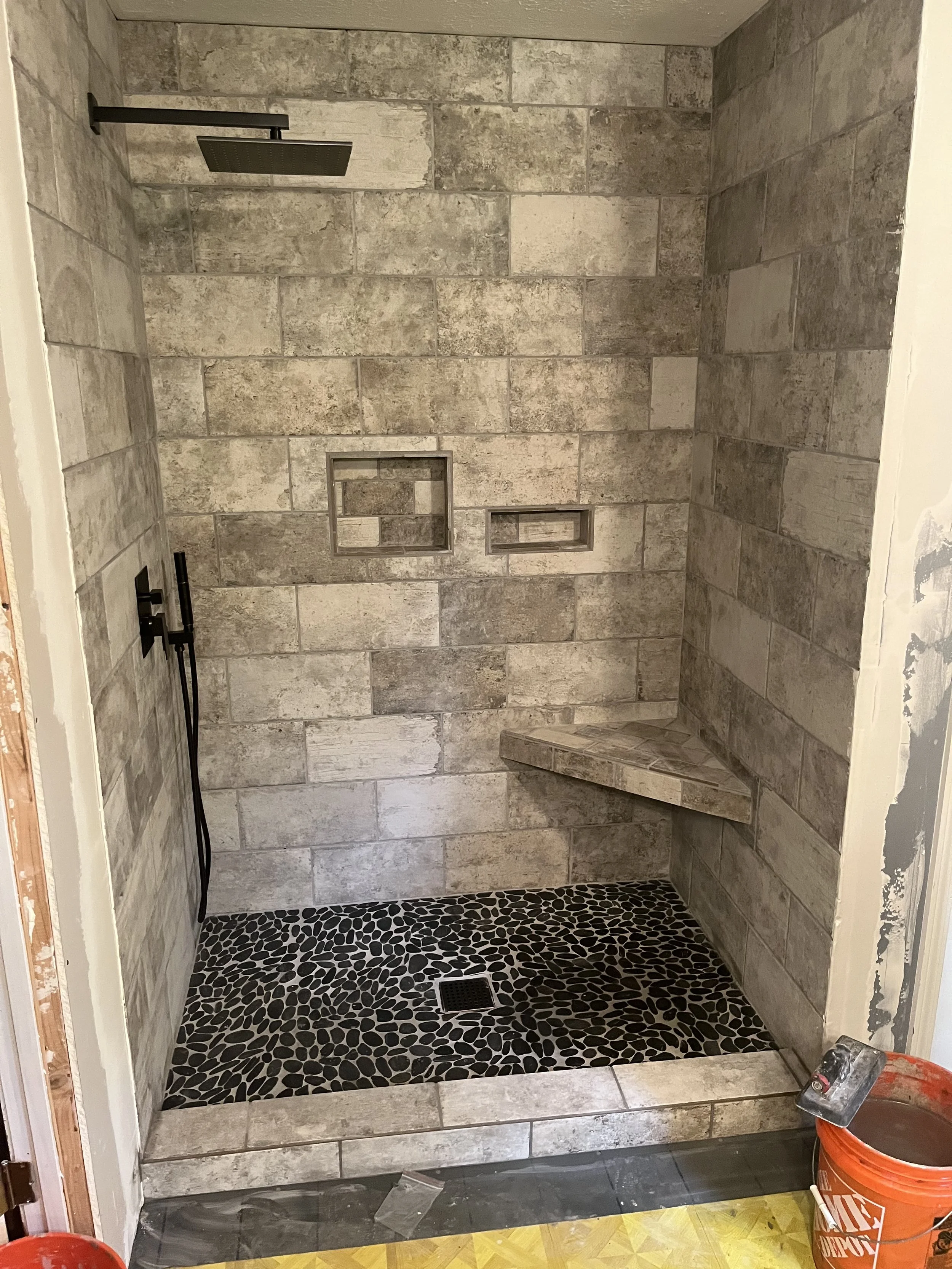 A modern shower with beige and gray tiled walls, a black rainfall showerhead, a handheld showerhead, two built-in shelving niches, a built-in corner bench, a black drain, and black pebble tile flooring.