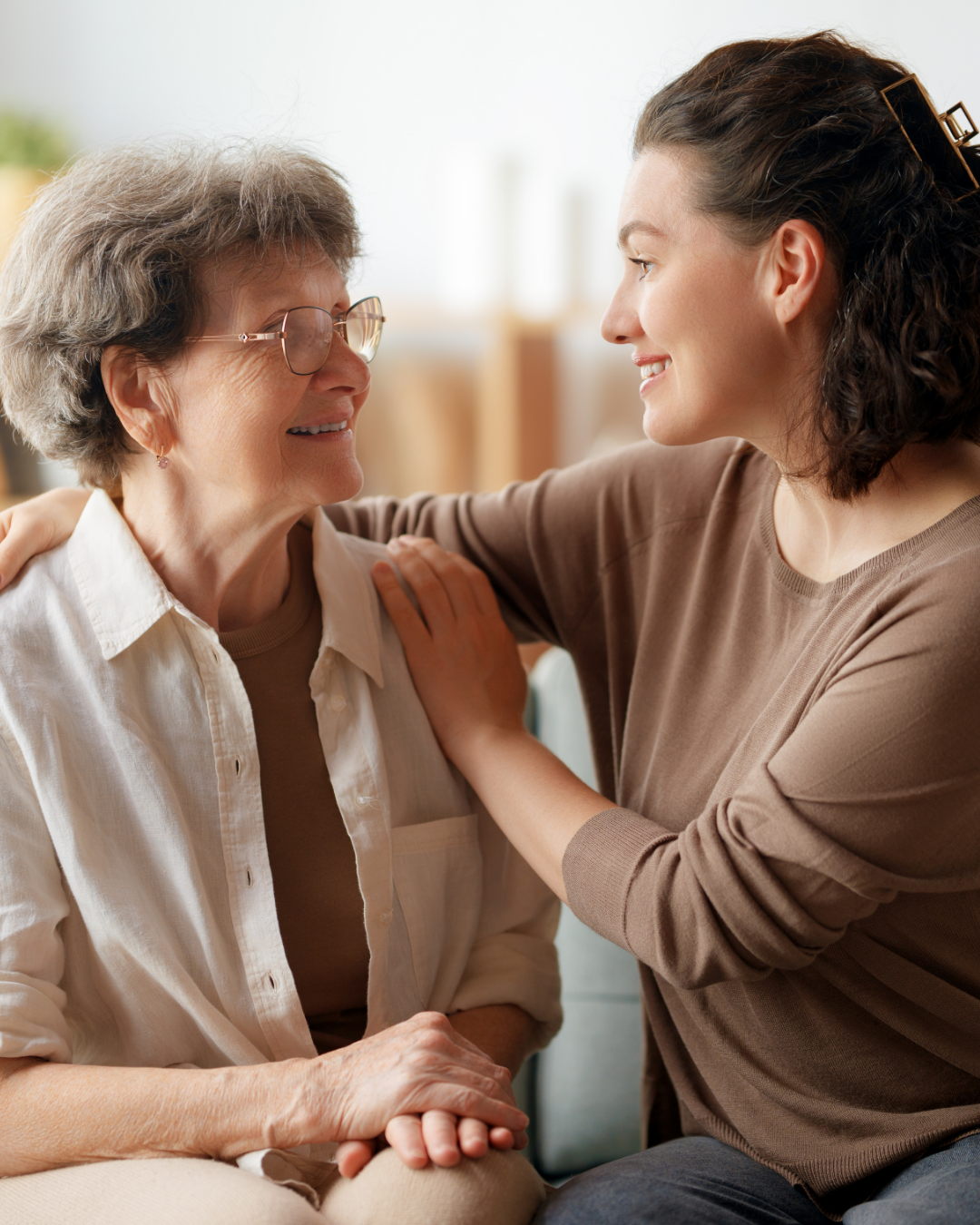 A young woman and an elderly woman smiling at each other, with the young woman placing her hand on the elderly woman's shoulder in a warm embrace.