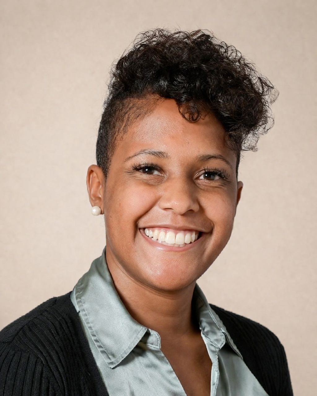 Close-up portrait of a smiling woman with short curly black hair, wearing pearl earrings, a light gray collared shirt, and a black blazer, against a plain beige background.