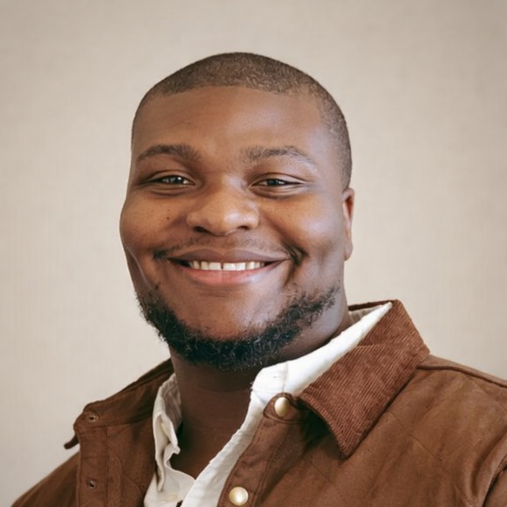 A smiling young man with short hair and a beard, wearing a white shirt and a brown jacket, standing against a plain background.