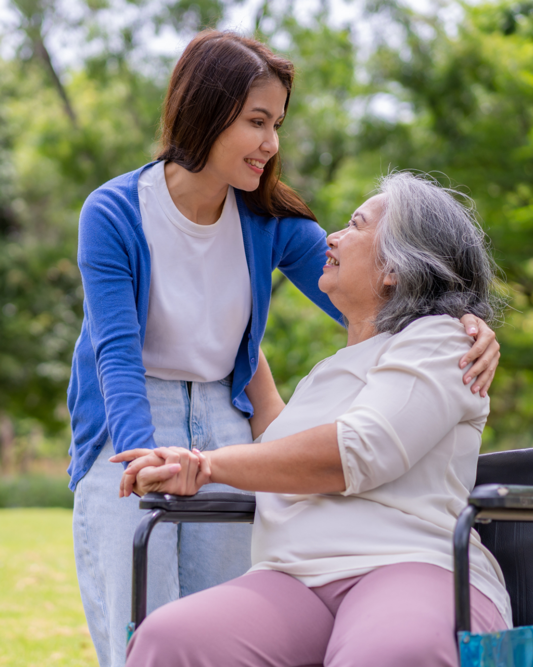 A young woman with brown hair smiling and leaning toward an elderly woman in a wheelchair, with a green outdoor park background.
