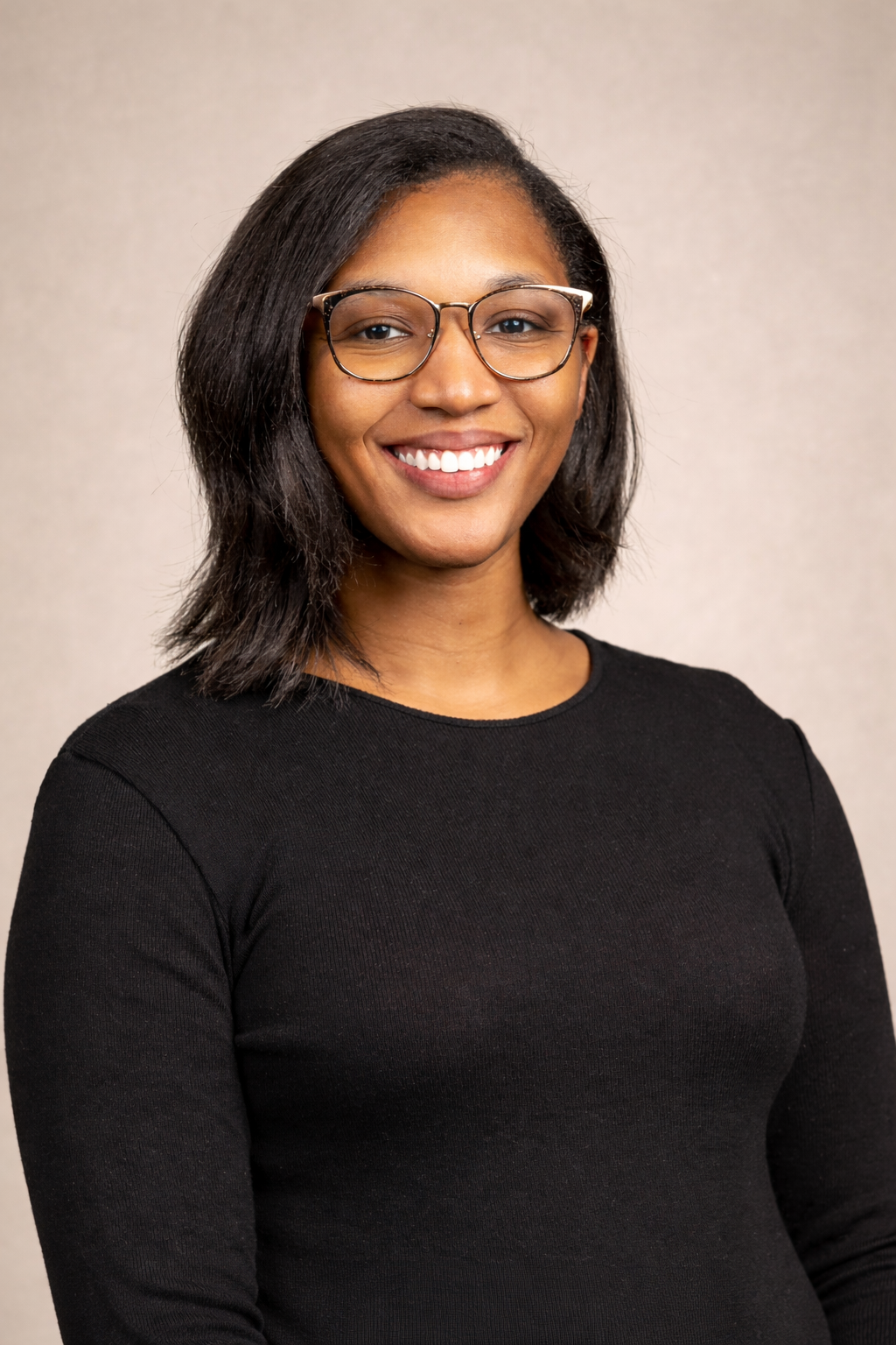 A young African American woman with shoulder-length black hair, wearing glasses and a black long-sleeve shirt, smiling at the camera against a plain light gray background.