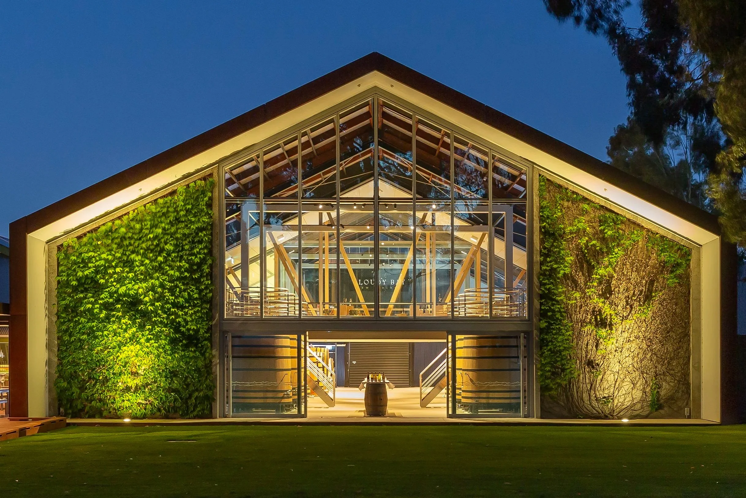 Night view of a modern building with glass front, wooden beams, and greenery on either side, illuminated with interior and exterior lights.