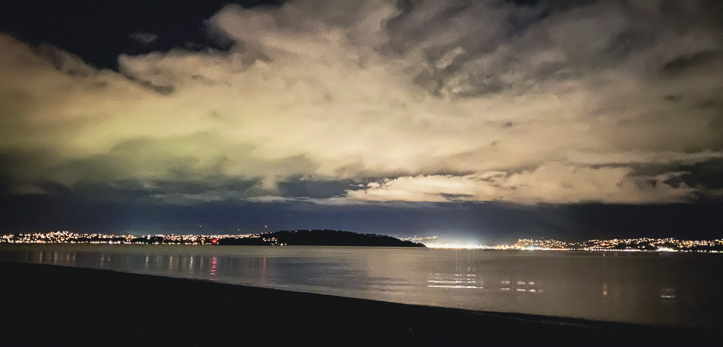 Nighttime landscape over a body of water with city lights in the distance, a cloudy sky illuminated by a distant storm, and a dark silhouette of land.