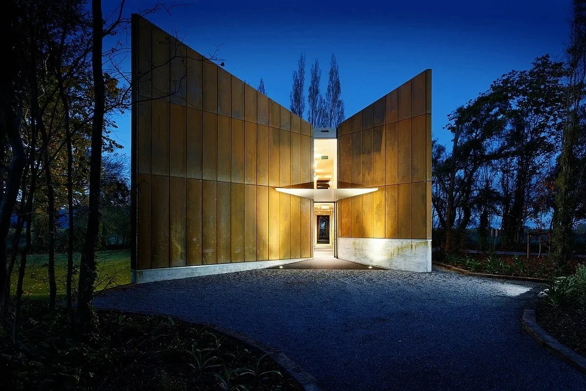 A modern building with large corten steel panels on each side, illuminated from within, located in a wooded area at dusk or night.