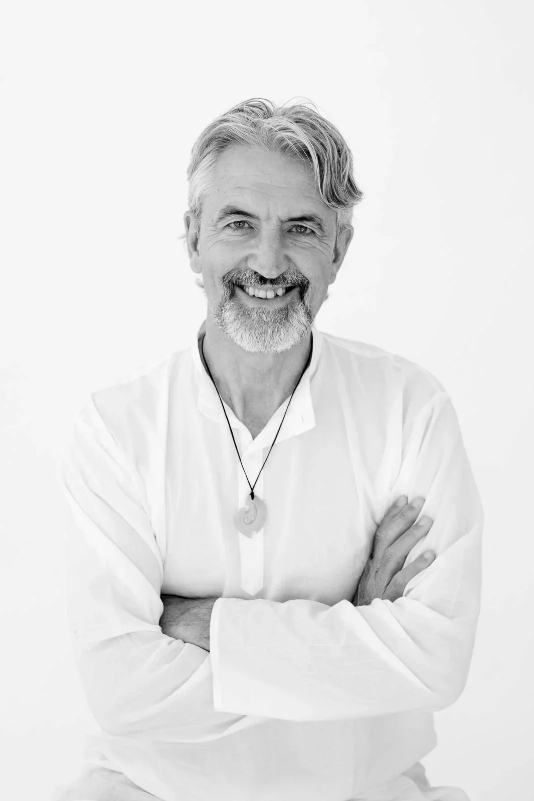 Black and white portrait of a smiling good looking middle-aged man with a beard, wearing a white shirt and cross-armed, against a plain light background.