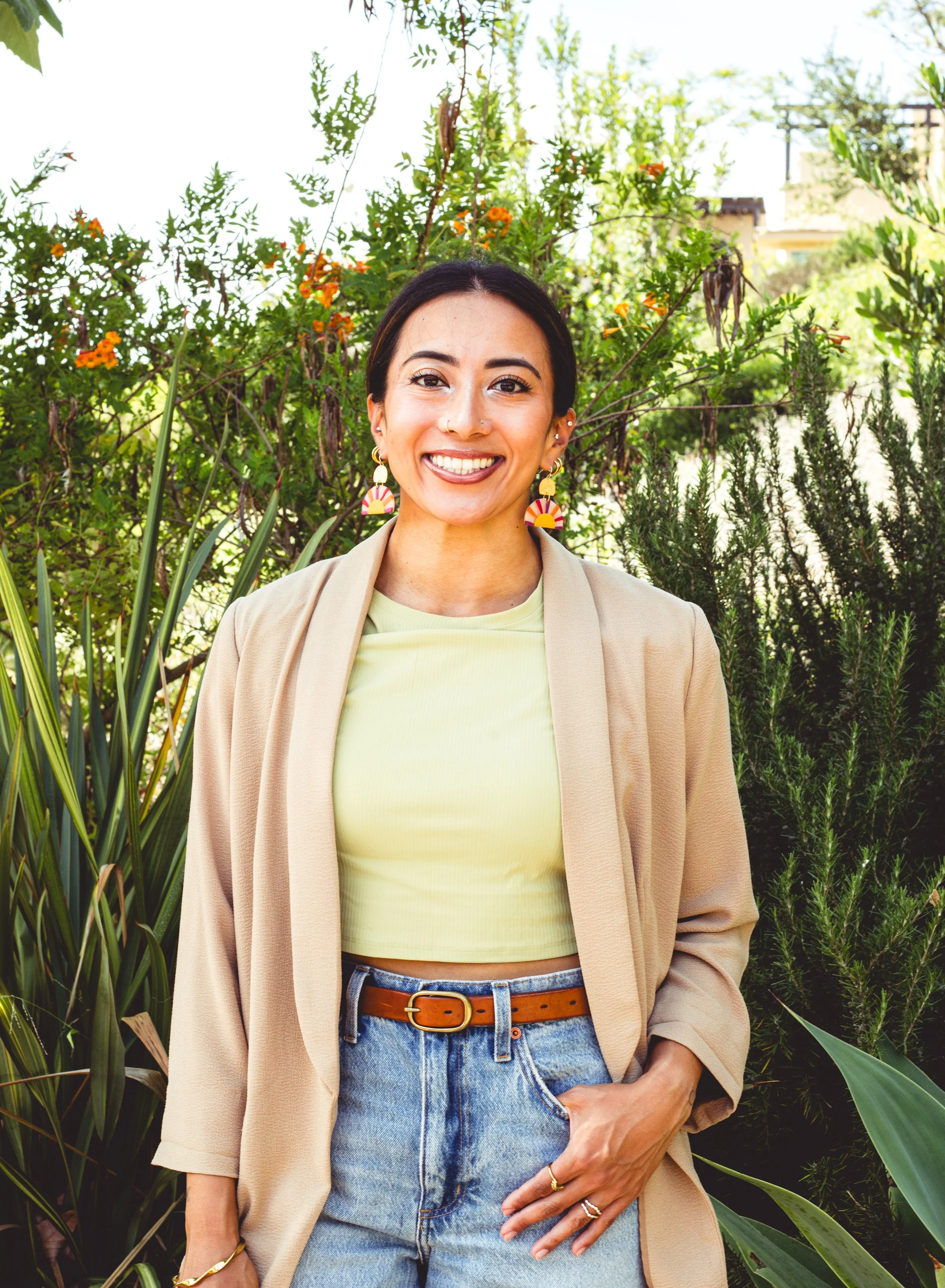 Rima Sheehab, an operations consultant and integrative health coach in San Diego, California, smiling outdoors in a beige blazer, lime green top, and blue jeans amidst lush green plants.