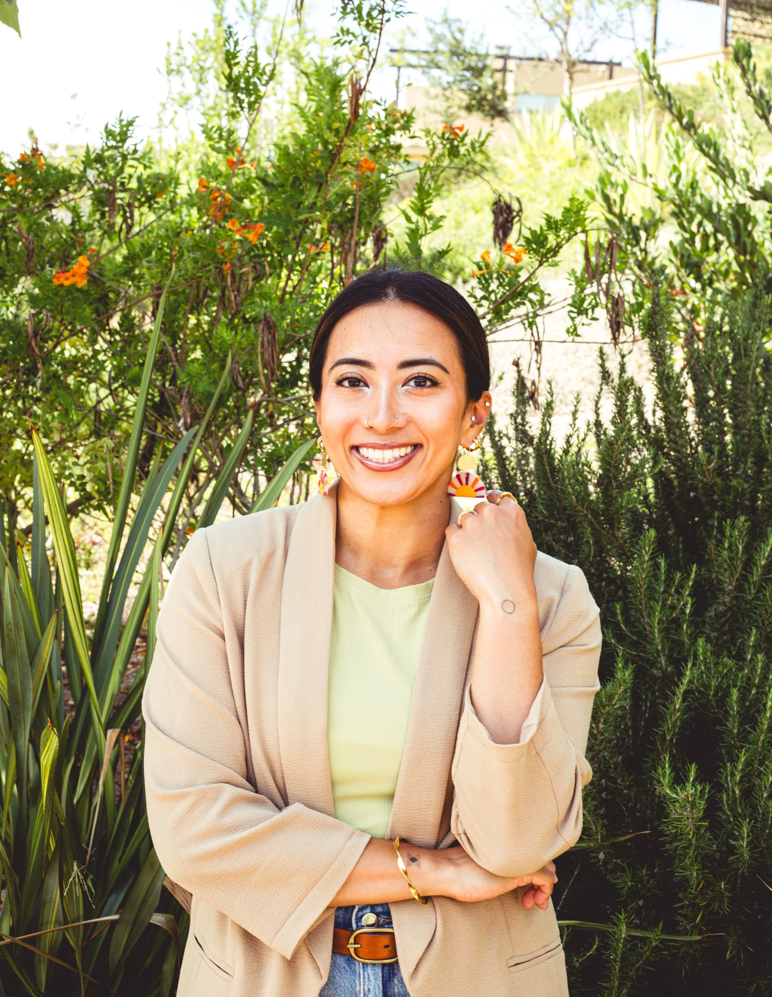 Headshot portrait of Rima Sheehab, founder of Healthy and Rising, wearing a beige blazer and colorful earrings while standing in front of vibrant green garden foliage in San Diego, California.