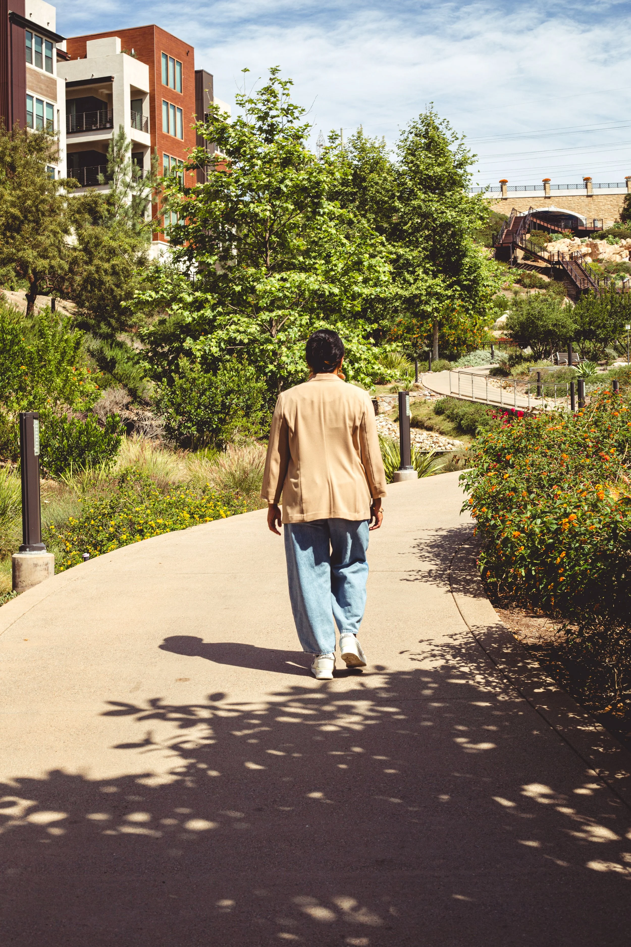 Rima Sheehab, a healthcare and tech operations consultant, wearing a beige jacket and jeans while walking down a concrete park pathway near modern urban buildings in San Diego, California.
