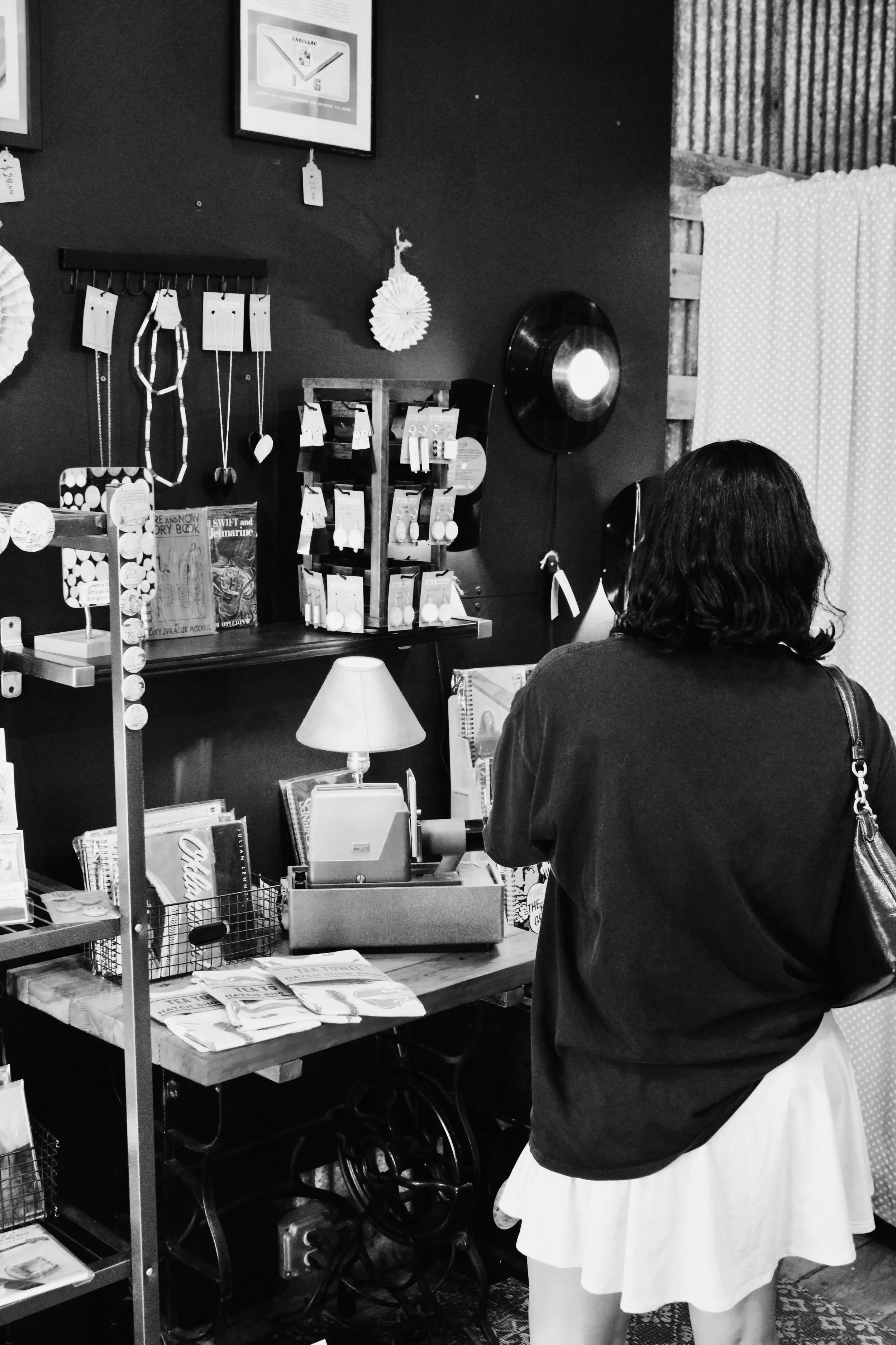 A woman standing at a small retail checkout counter in a store, looking away from the camera. The counter has a cash register, a small lamp, and various magazines and items on display. The background features jewelry and greeting cards on the wall.