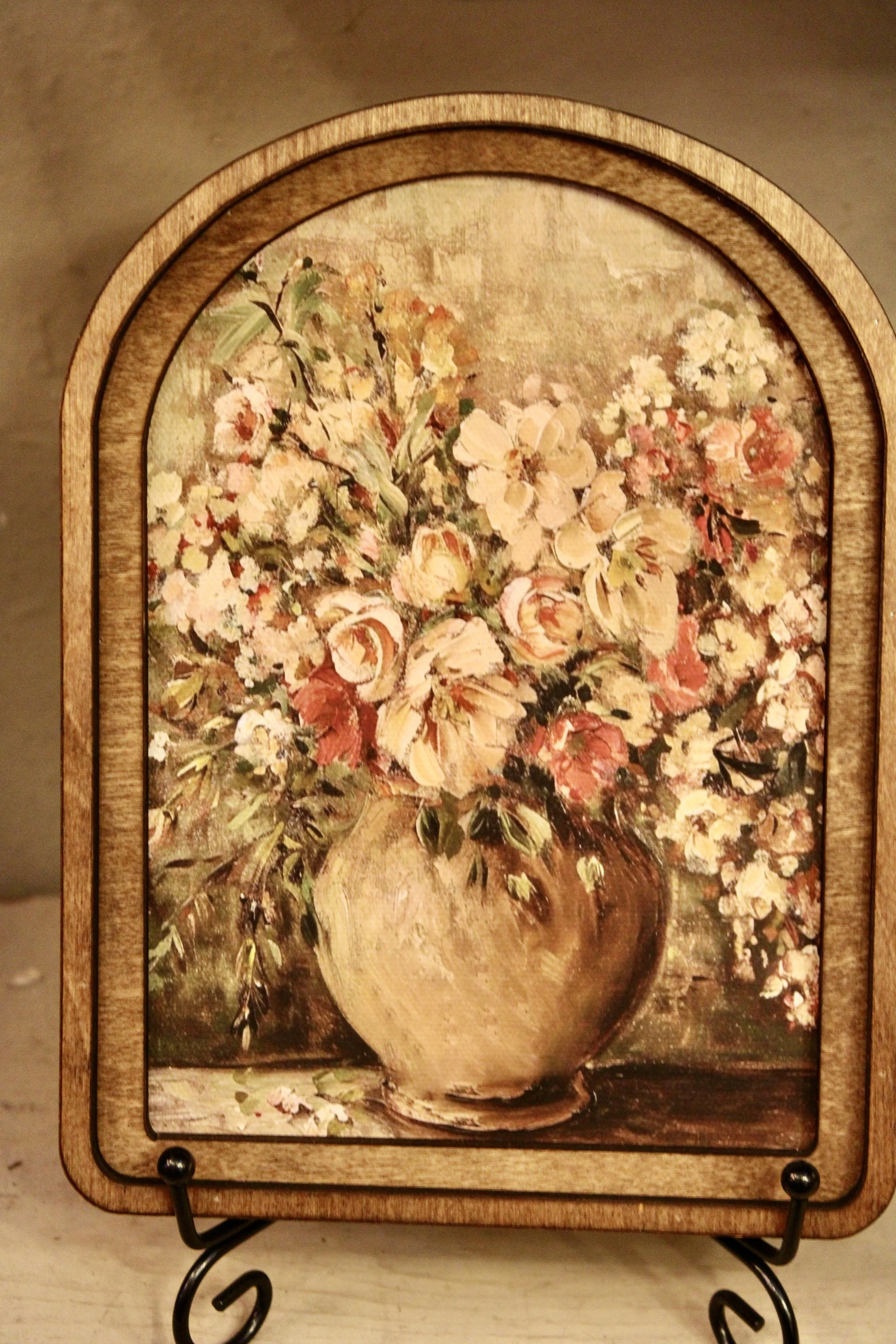A framed painting of a large bouquet of flowers in a vase, placed on a table. The flowers are in soft pastel shades of pink, cream, and white, with green leaves.
