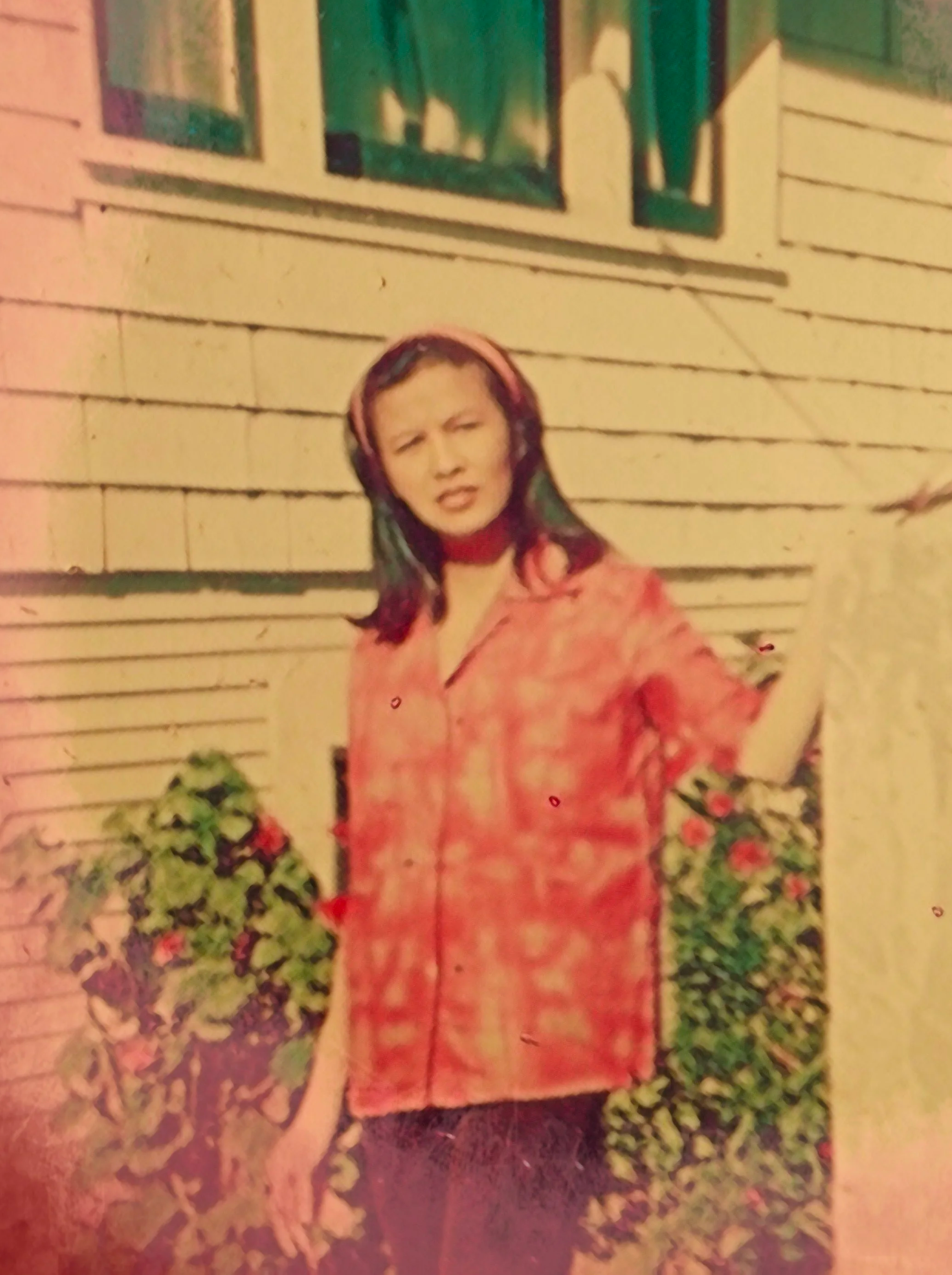 A woman wearing a pink top and pink headband standing outside near a yellow house with green-framed windows and surrounded by plants and flowers.