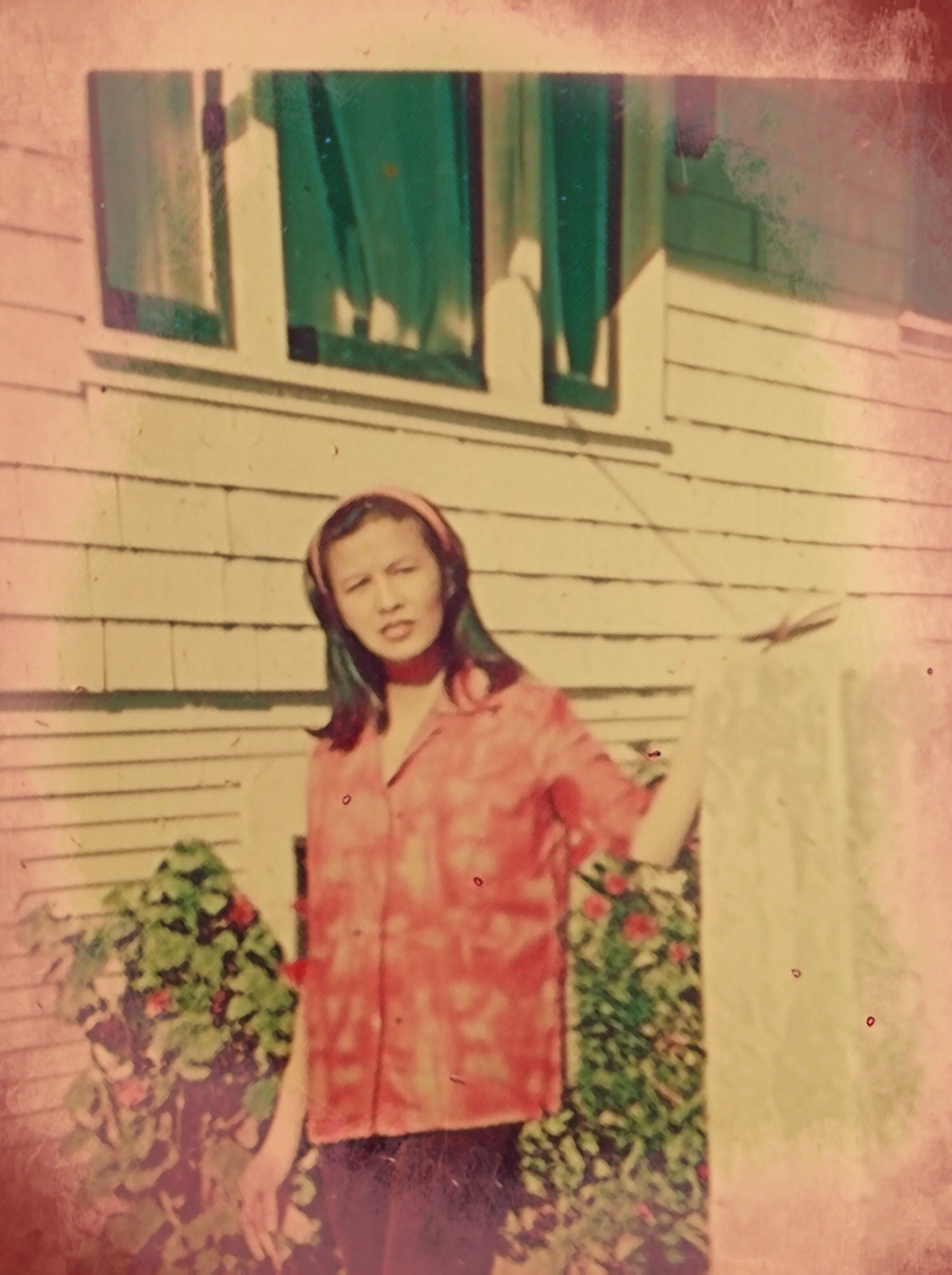 A woman standing in front of a house with white siding, surrounded by green plants and flowers.