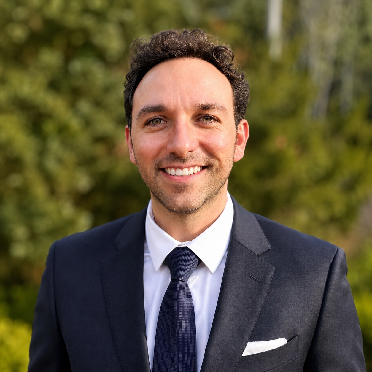 Daniel Guzman, founder of Prymax Advisory, smiling in a business suit outdoors with a blurred green background