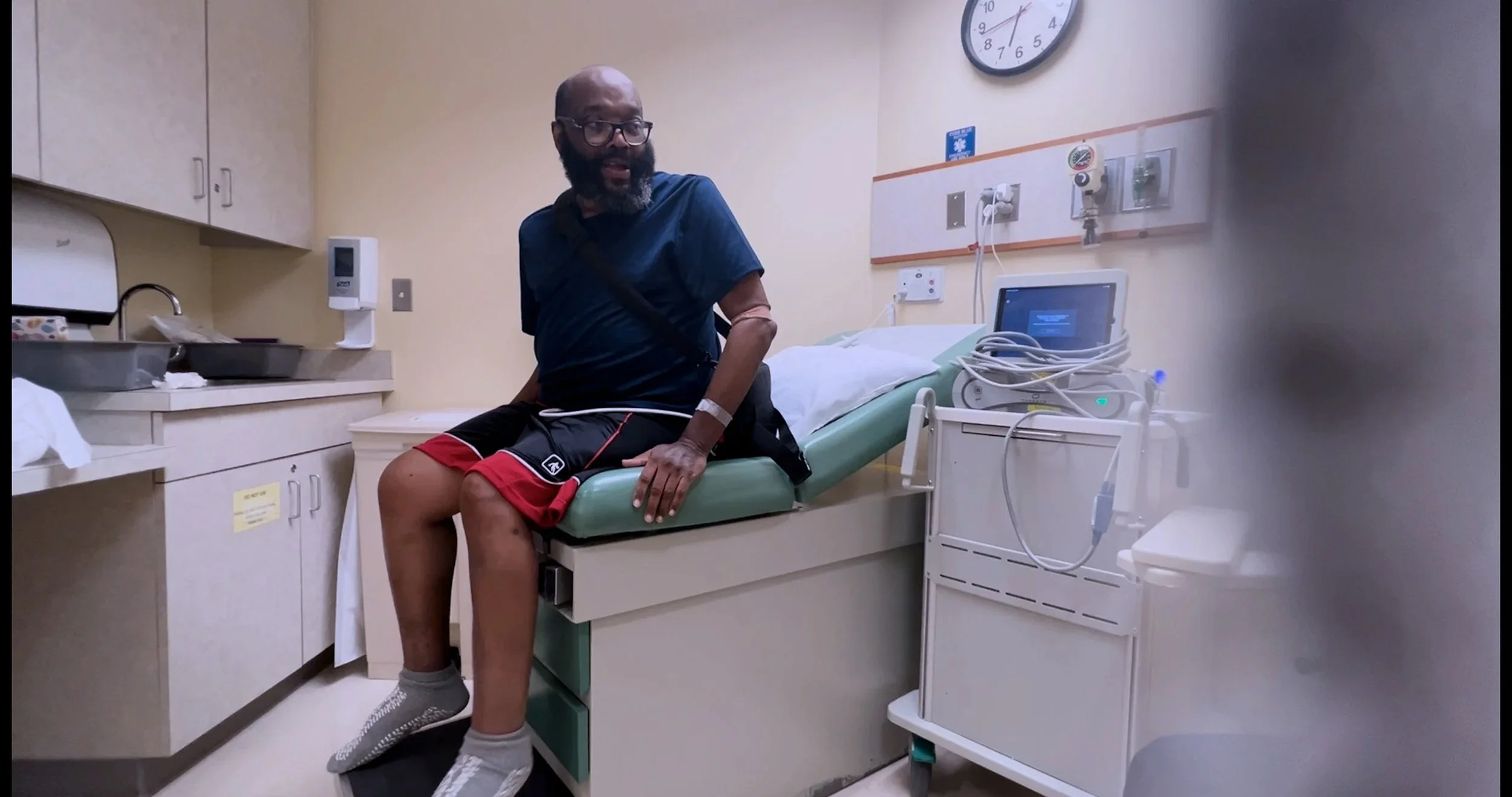 A man sitting on the edge of a hospital bed in a medical examination room, wearing casual clothes and hospital wristband, with medical equipment and cabinets around him.