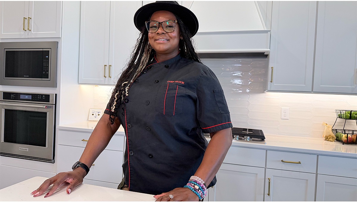 A woman wearing a chef's uniform and hat, smiling in a modern white kitchen with stainless steel appliances.