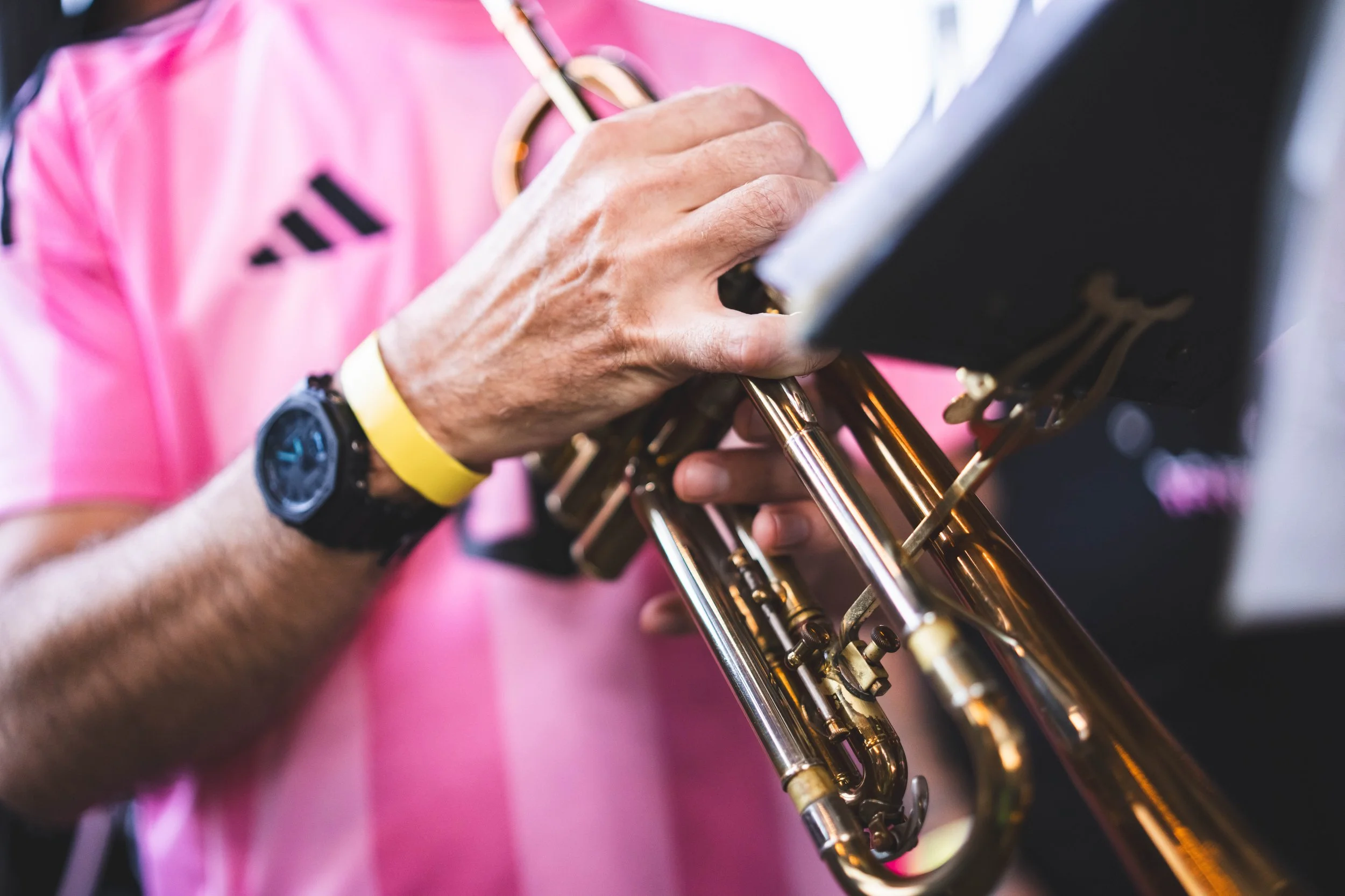 A person holding a trumpet with a music stand nearby, wearing a pink shirt and a black watch.
