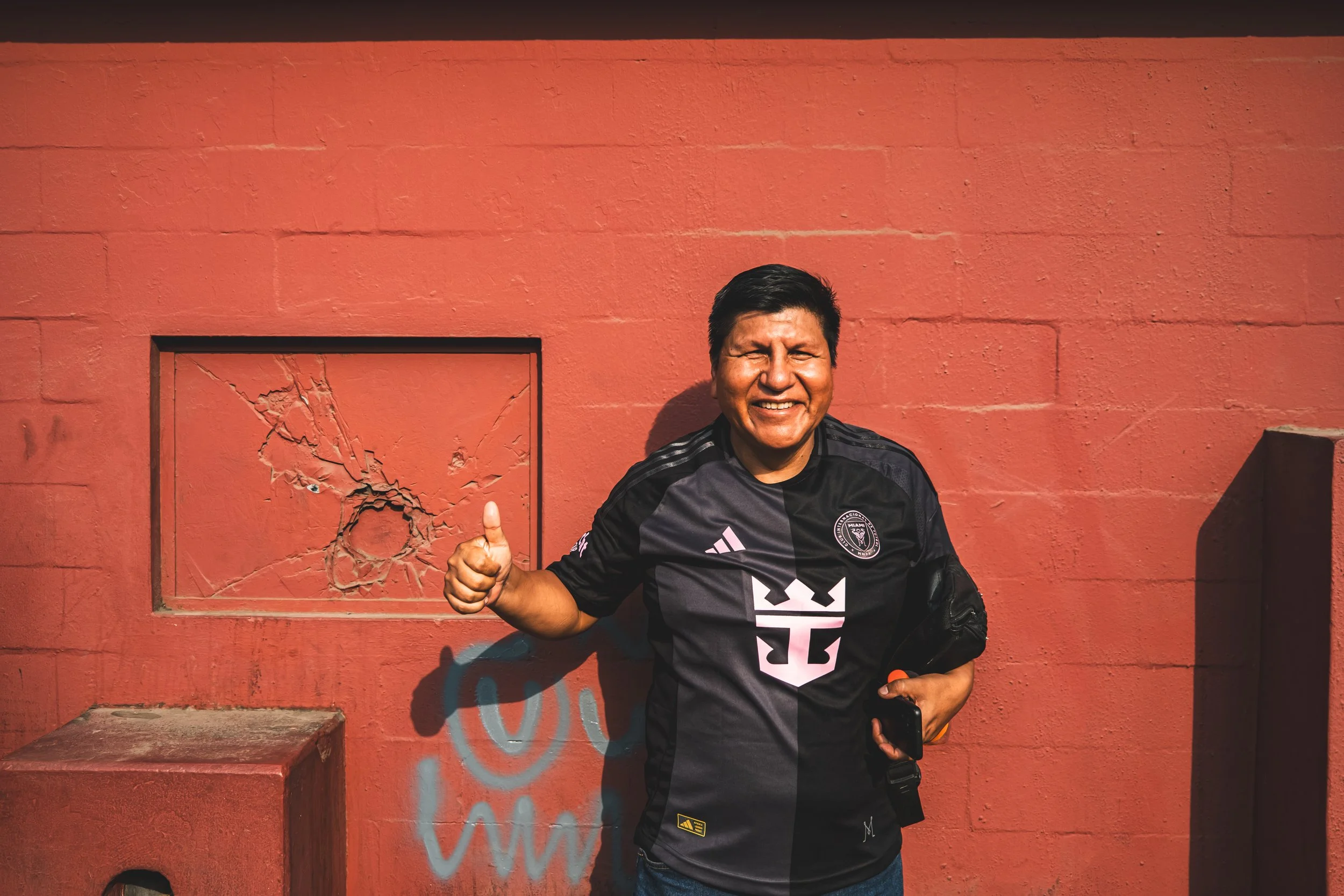A man smiling and giving a thumbs-up while standing in front of a red wall with a broken window.