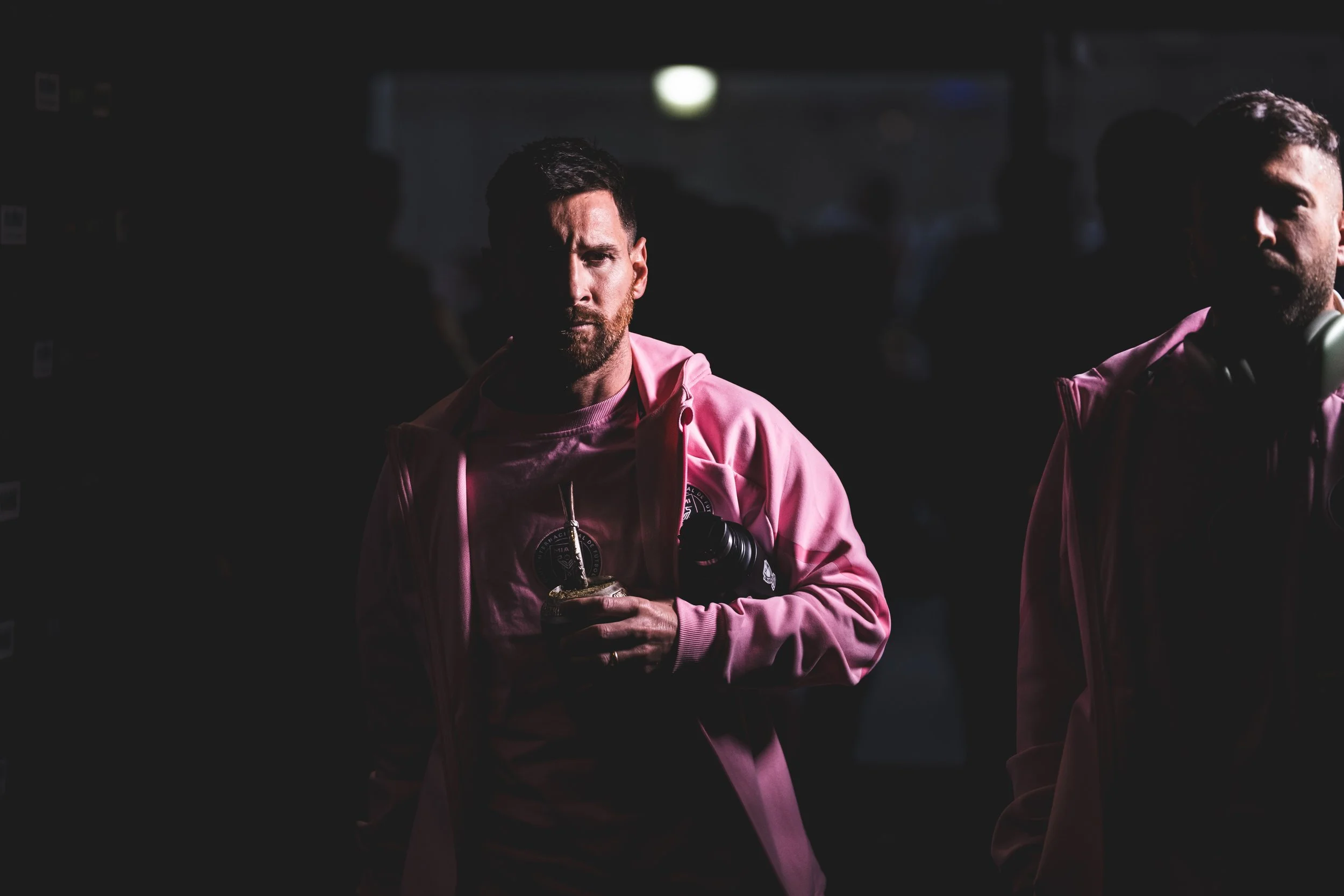 A man with a beard wearing a pink hoodie, holding a drink and a camera, standing in a dark environment with other people in the background.