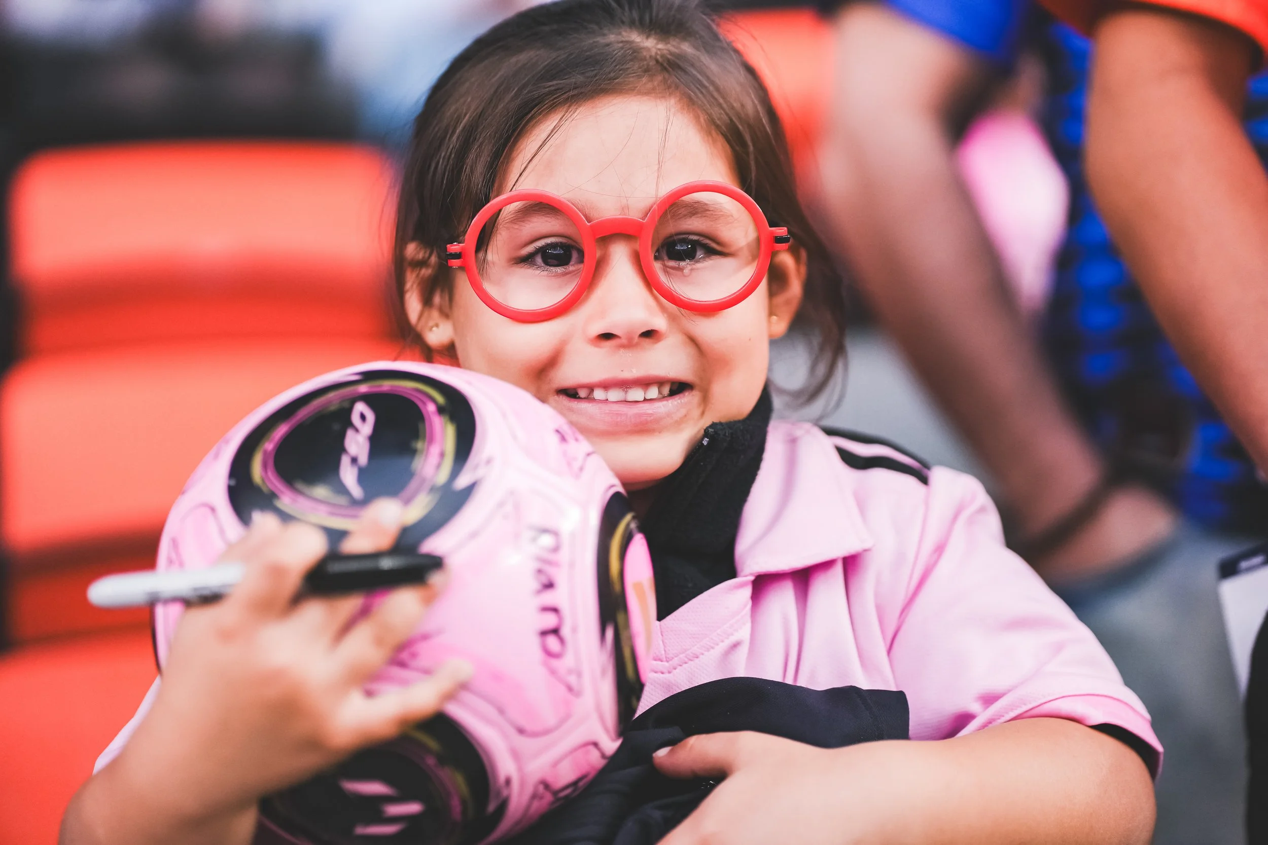 Young girl wearing red glasses holding a pink motorcycle helmet and a marker, smiling.