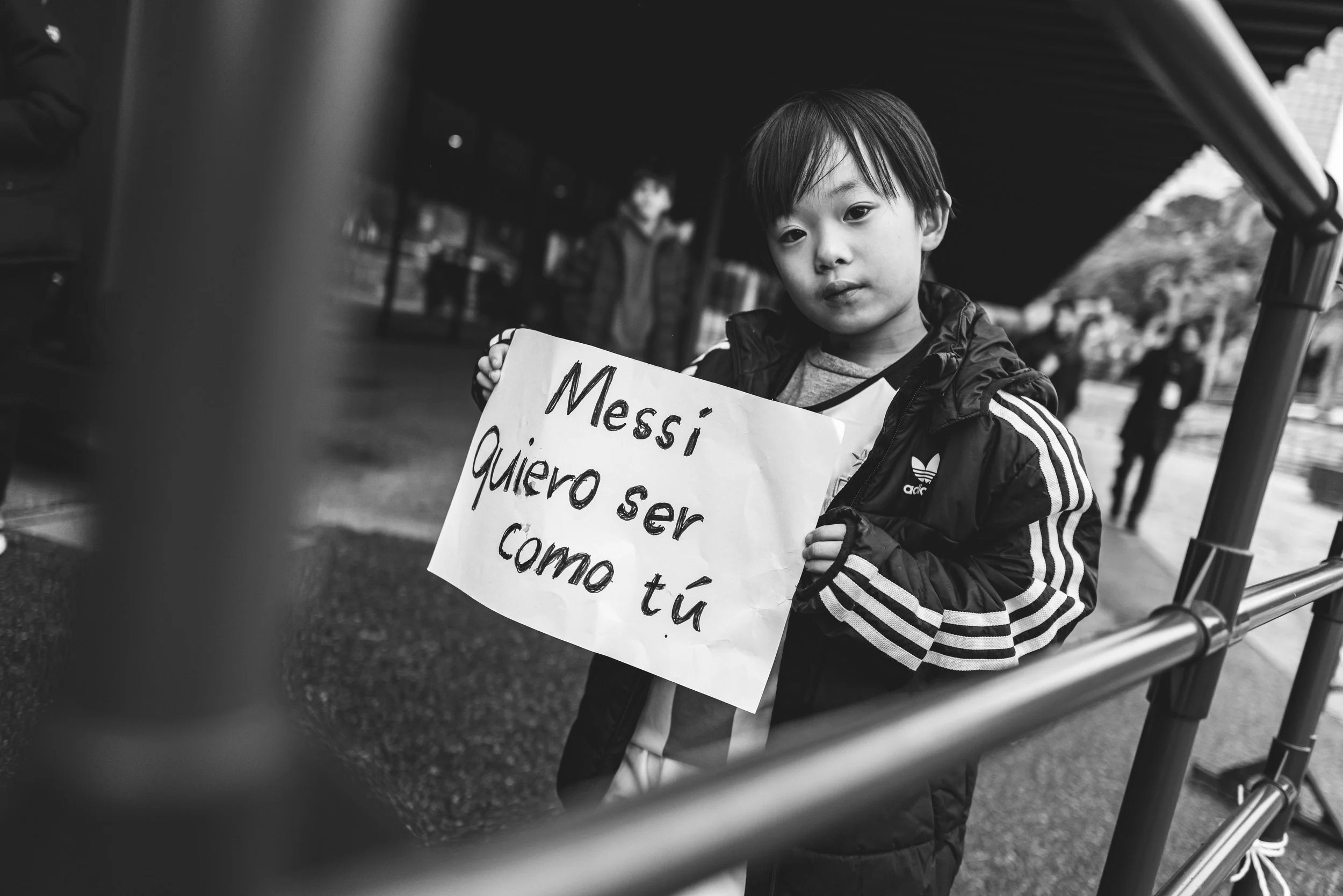 A young boy holding a handwritten sign in Spanish that says, 'Messi quiero ser como tú,' standing outdoors near a railing with a few people in the background.