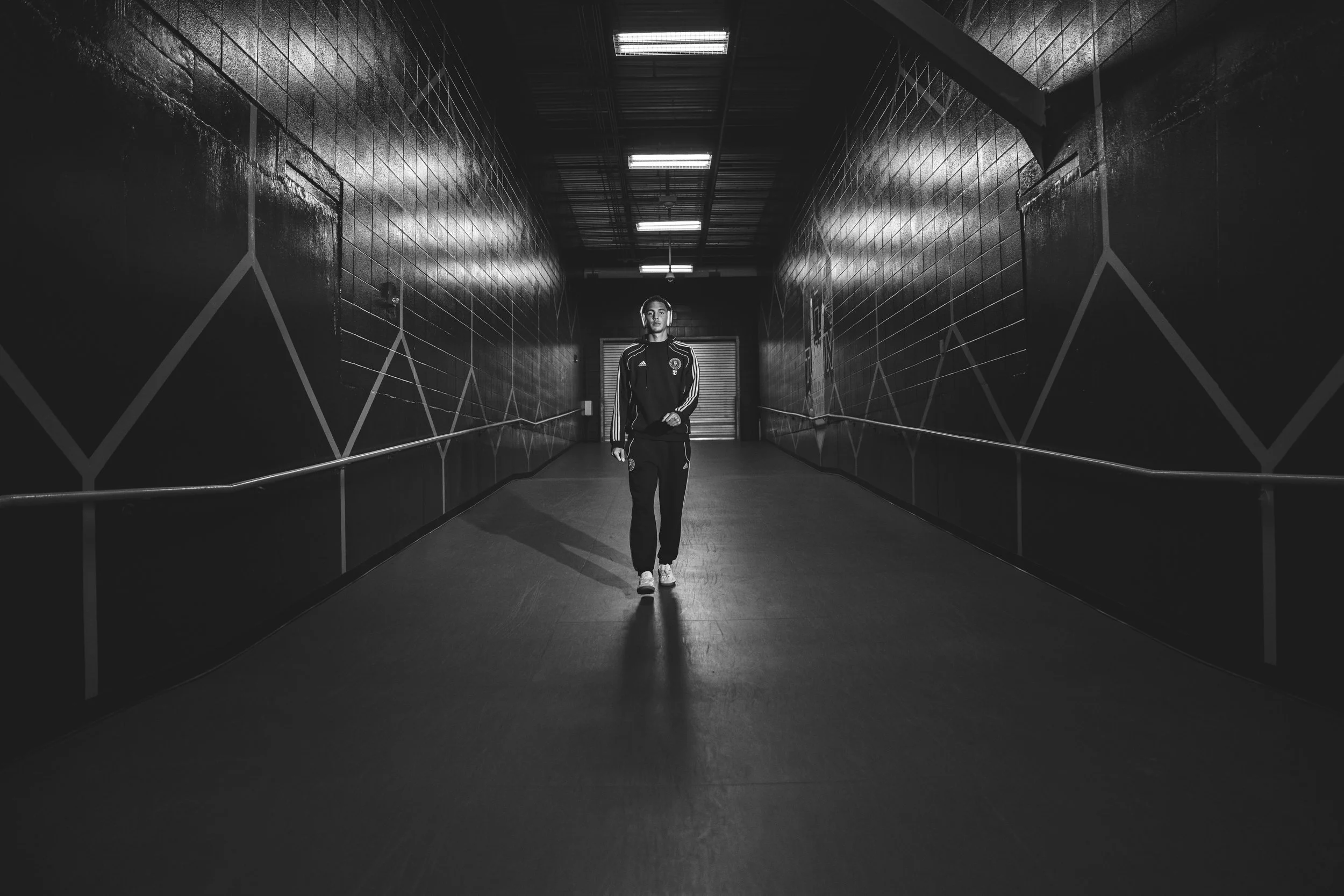 A person walking in a dimly lit underground tunnel with dark walls and geometric patterns, wearing sportswear.
