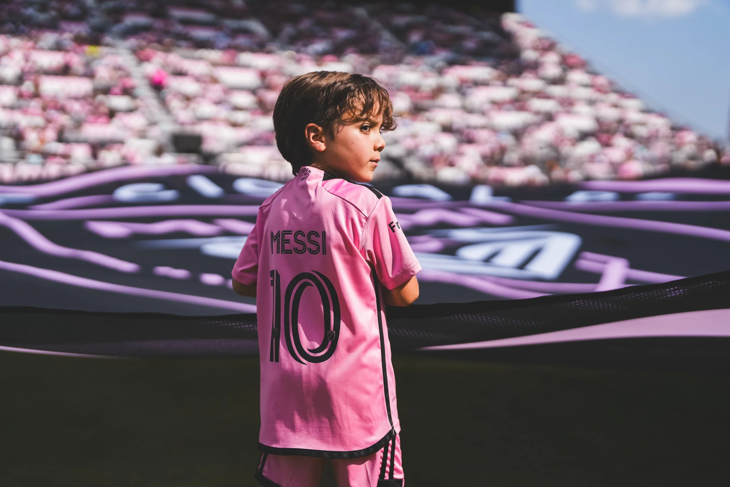 A young boy dressed in a pink soccer jersey with the name 'Messi' and the number 10 on the back, standing on a soccer field during a game or event.