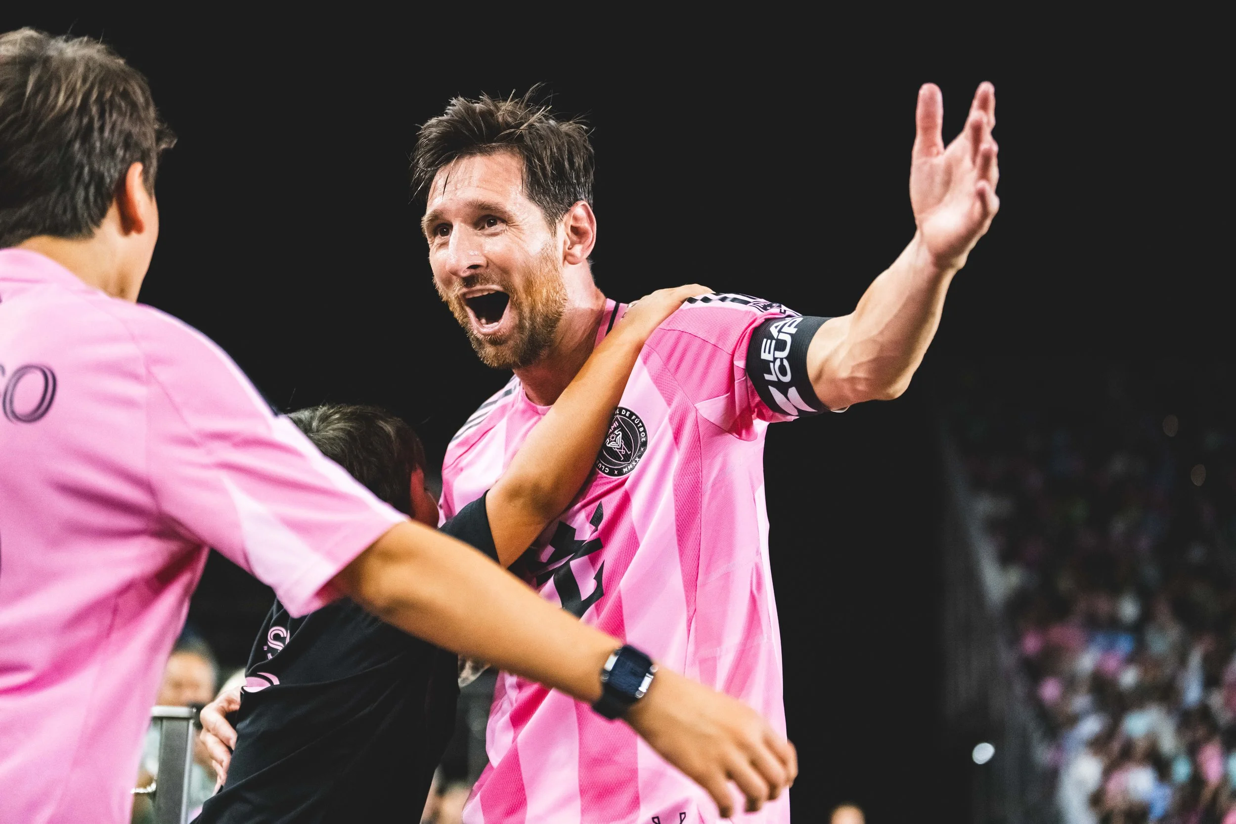 A man with a beard, wearing a pink sports jersey, celebrating with arms outstretched on a soccer field at night, with fans in the background.