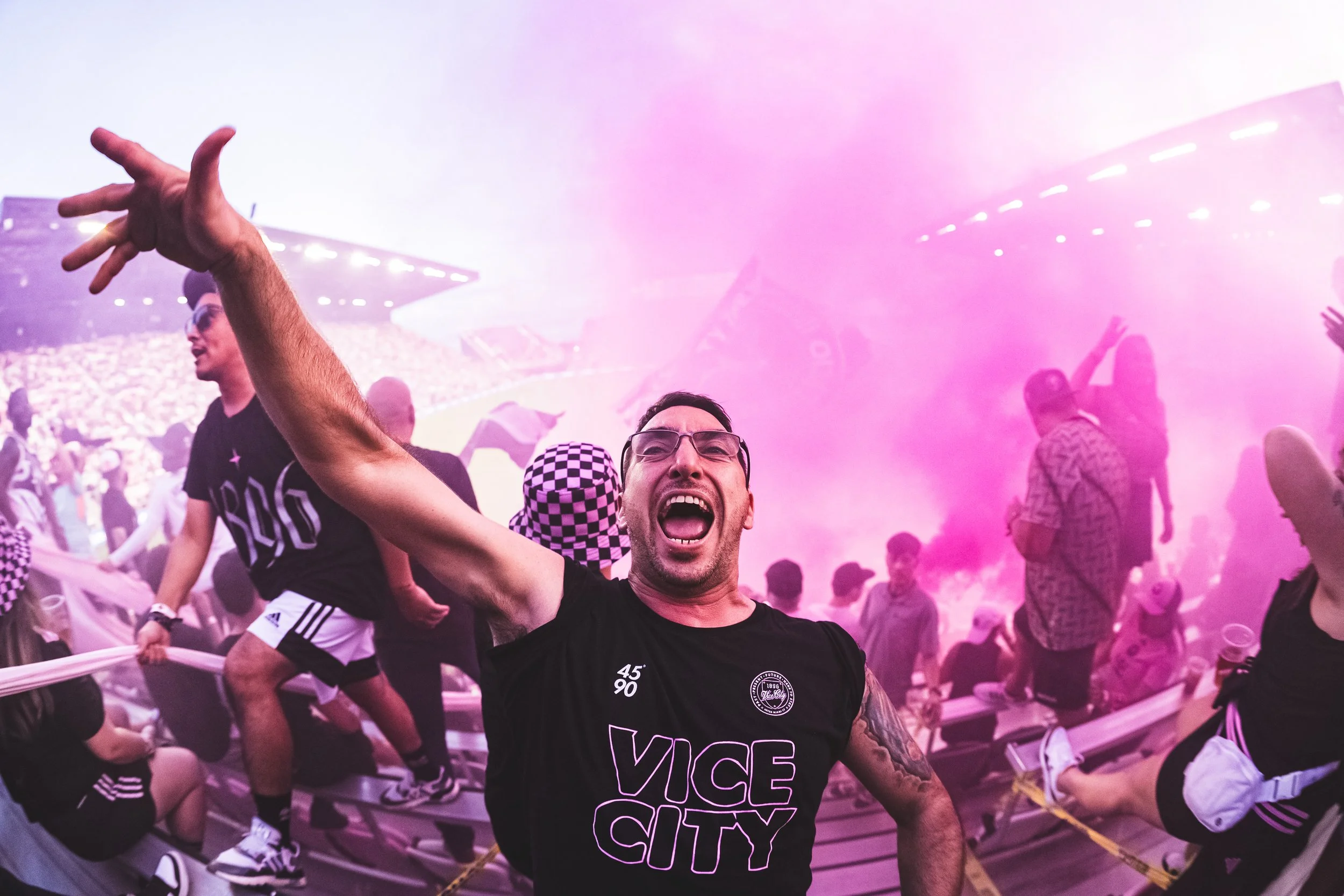 Crowd at a concert with pink stage lighting, with a man in the foreground raising his arm and shouting, wearing a black t-shirt that says "VICE CITY."