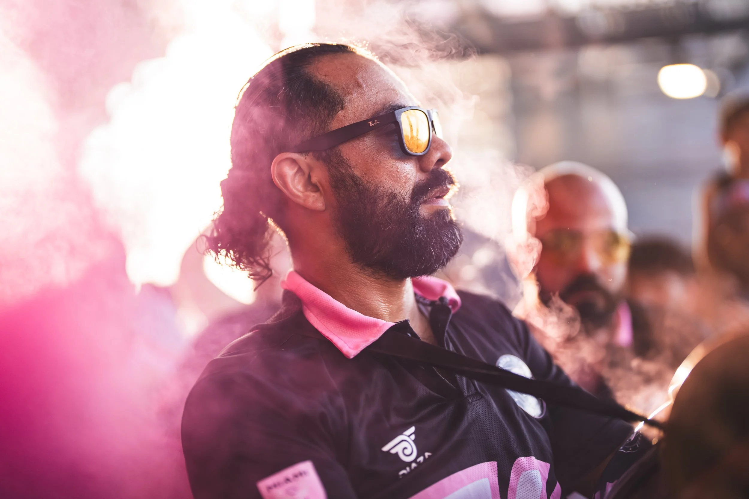 A man with a beard and long curly hair wearing sunglasses and a black sports jersey with pink accents, exhaling smoke at sunset or sunrise.
