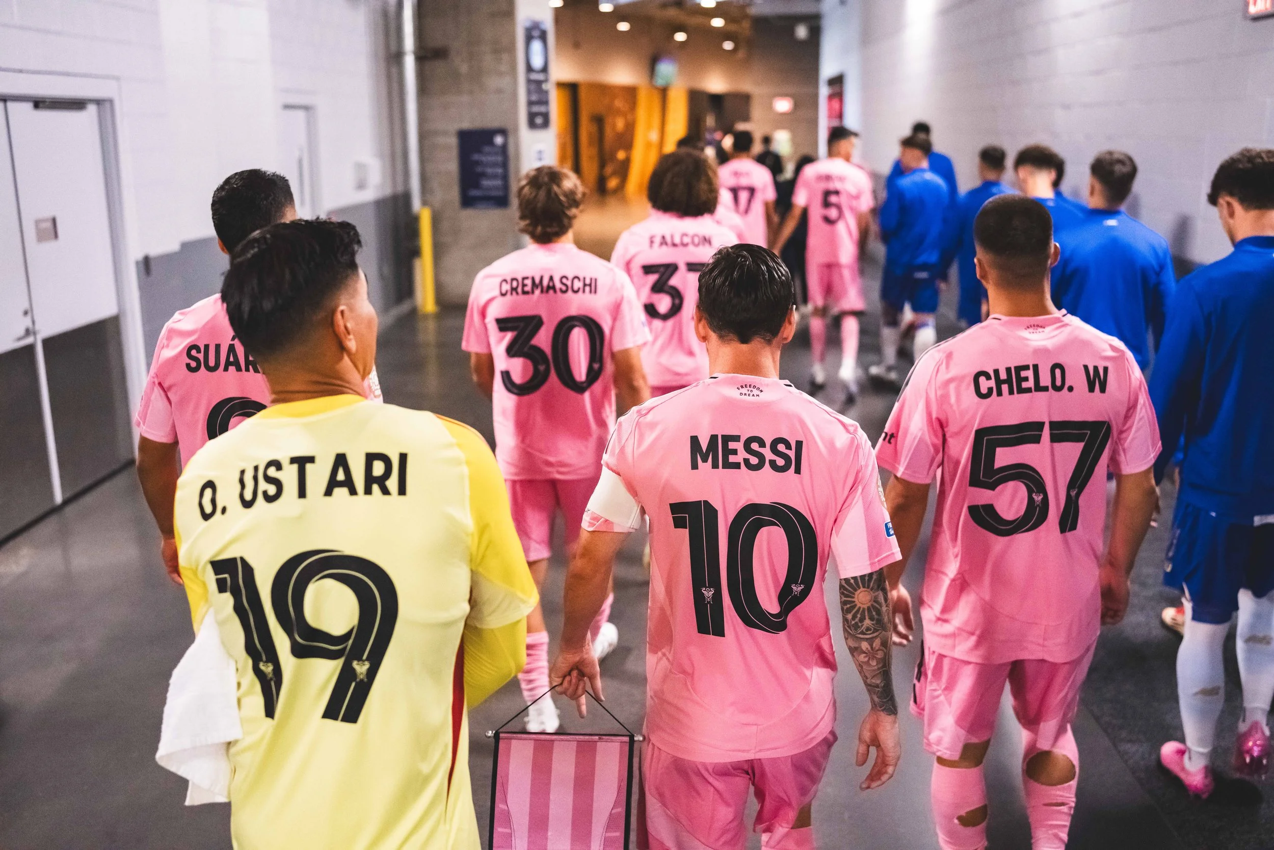 Soccer players walking in a locker room tunnel, wearing pink uniforms, with one player in yellow. The players' jerseys display their names and numbers, including Messi (10) and O. Ustari (19).