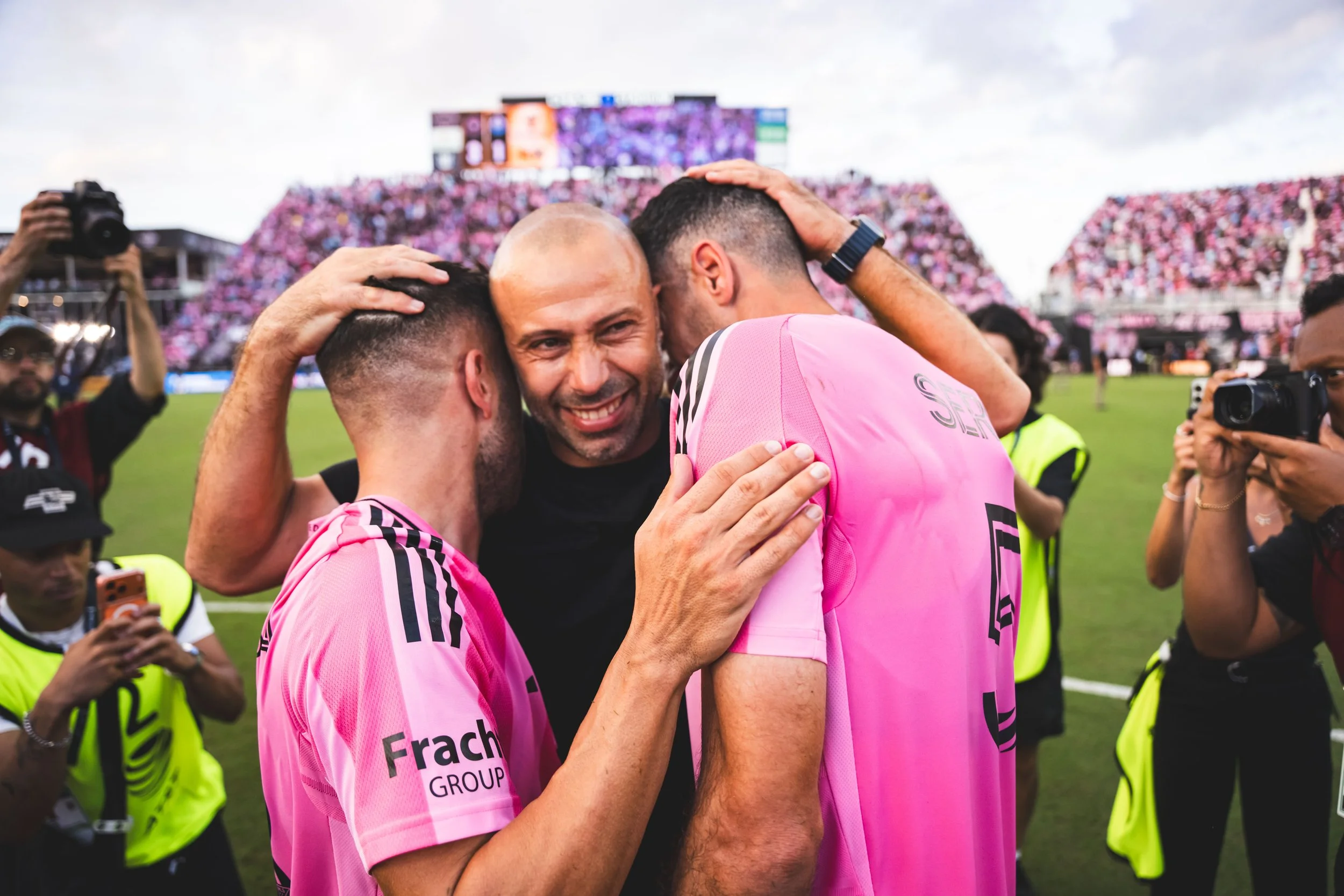 A team celebrates a victory on a soccer field, with two players and a man hugging, in a pink uniform, and numerous spectators and photographers in the background.