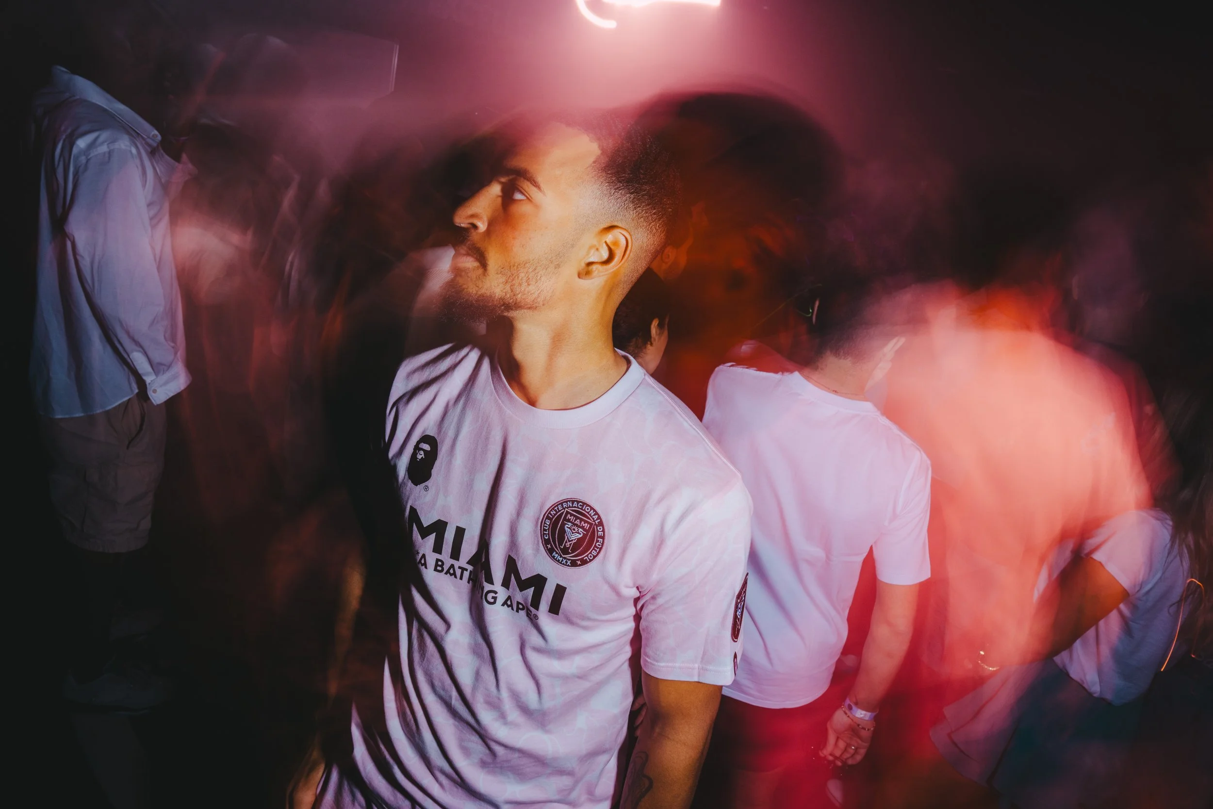 A young man with short hair and a beard wearing a white Miami football club T-shirt, standing in a dimly lit, crowded space with pink and orange lighting, and several other people around him.
