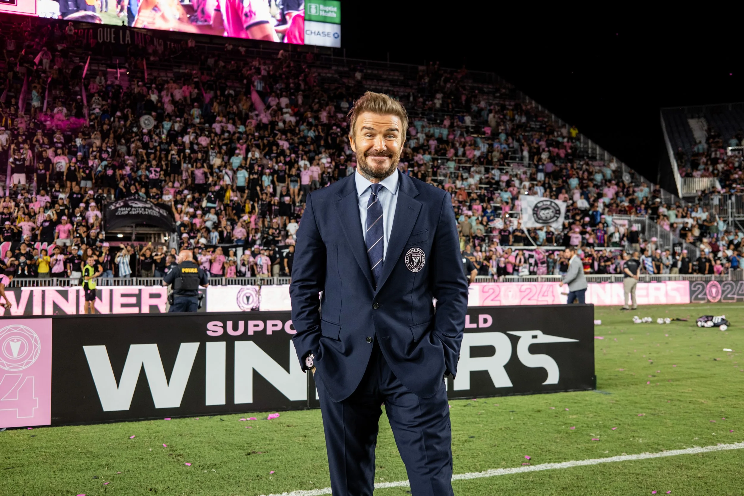 Man in a navy blue suit and tie standing on a sports field with a large crowd in the background, celebrating a victory at a sports event.