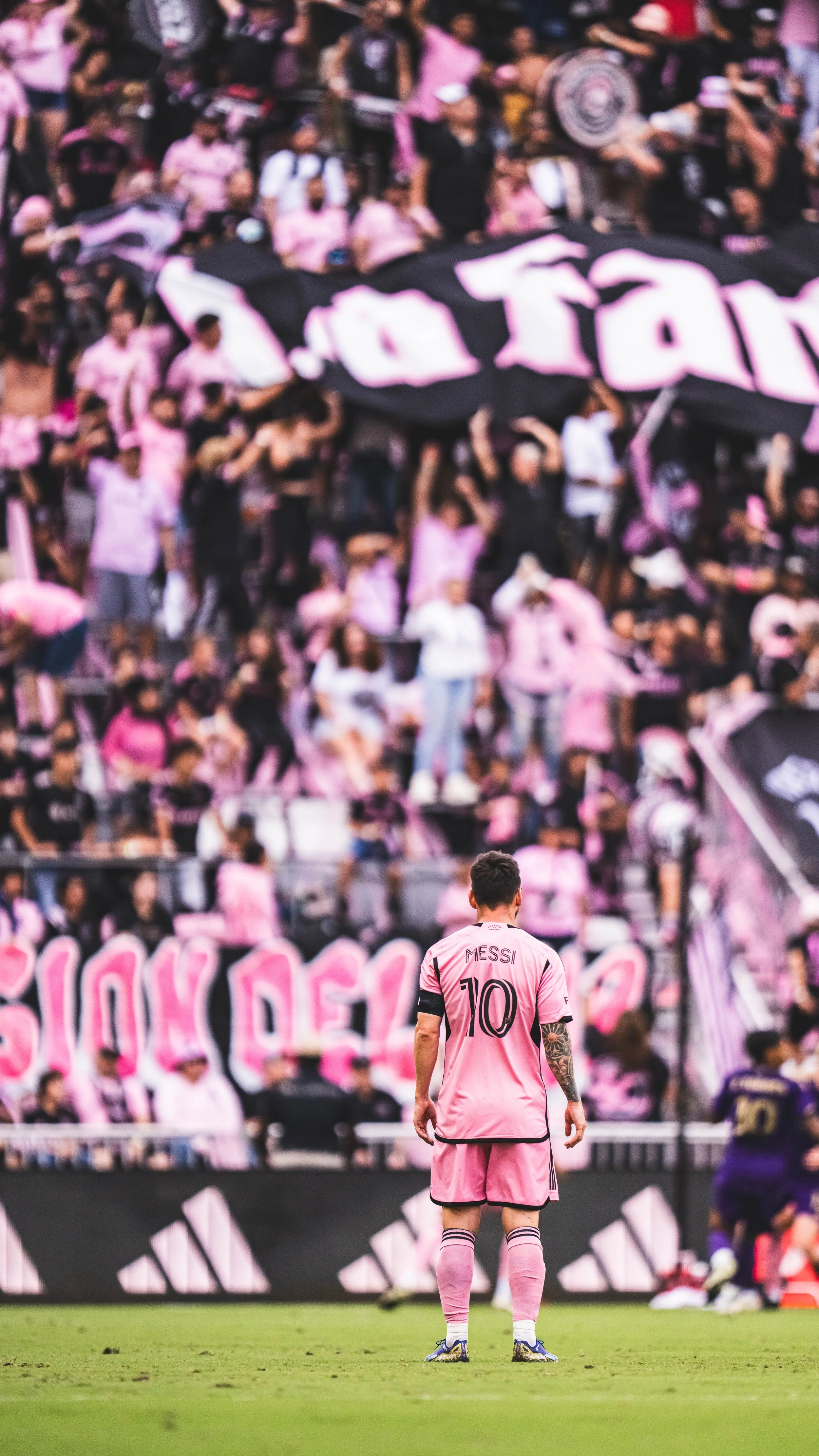 Soccer player Lionel Messi in pink Barcelona uniform standing on the field with a crowd of fans in pink and black jerseys behind him.