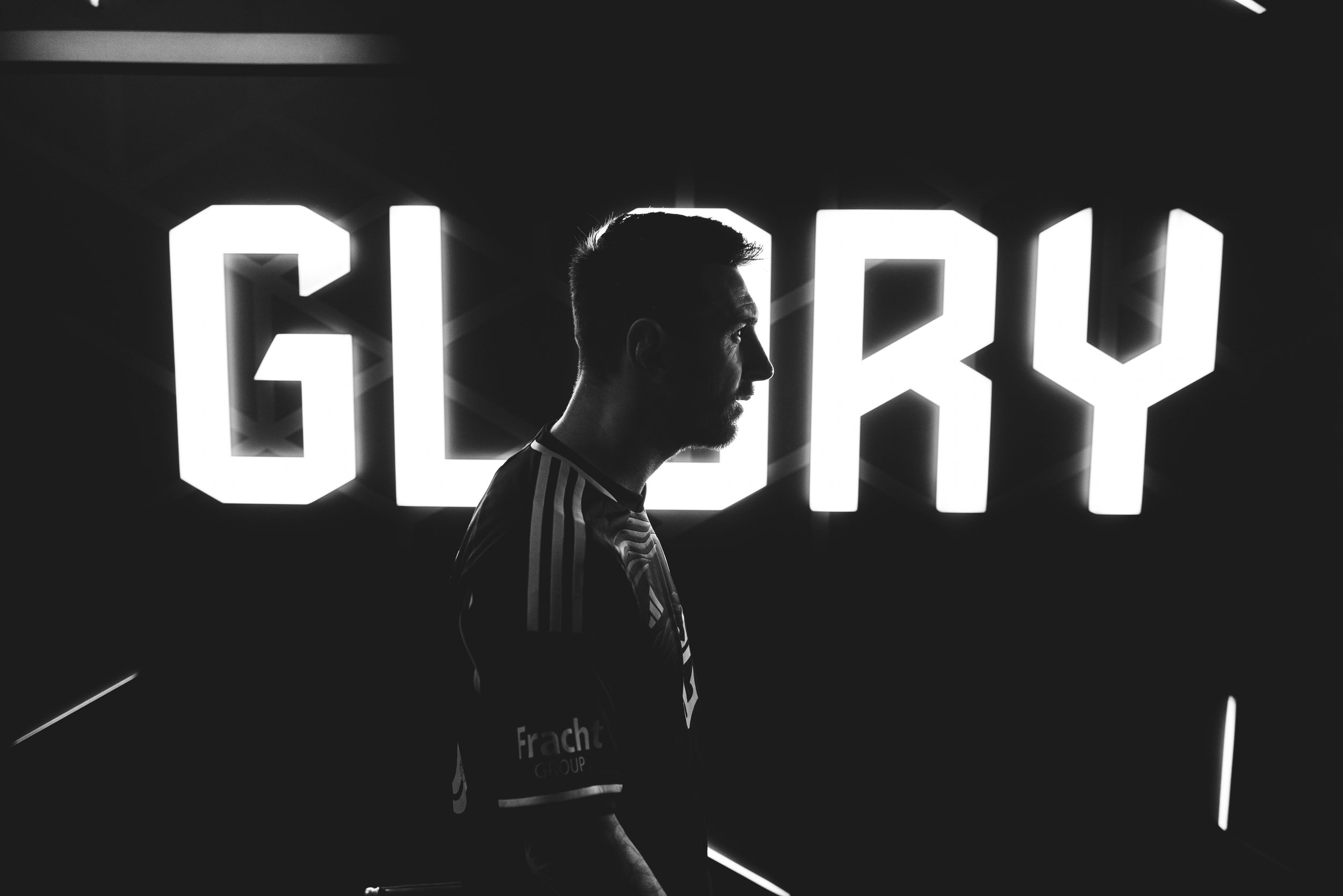 A man in profile standing in front of a bright sign that says 'GELRY' in bold illuminated letters, creating a silhouette effect.