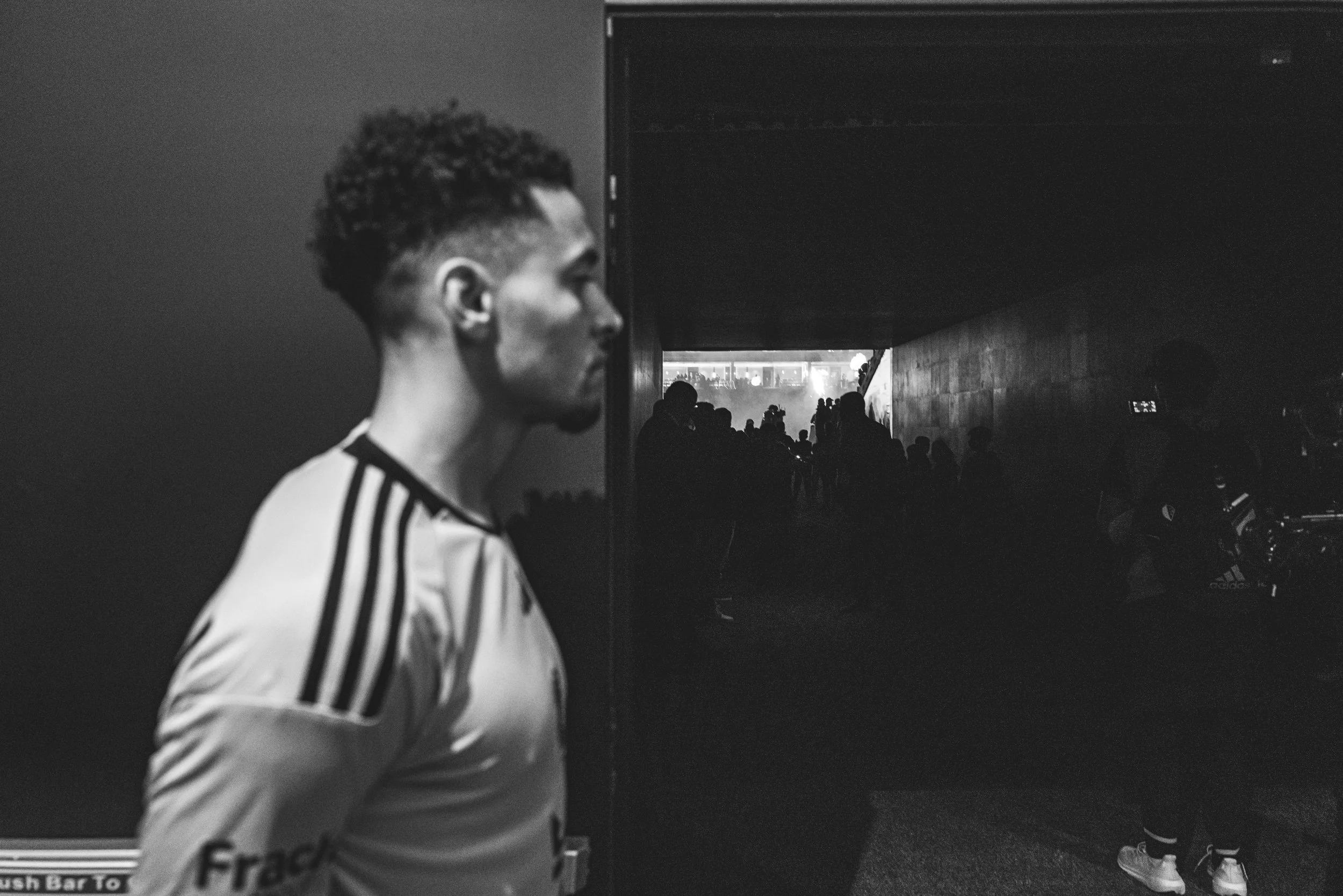 A black and white photo of a young man with curly hair and a beard, wearing a sports jersey, standing in a dark hallway looking to the side, with a crowd or group of people in the background.